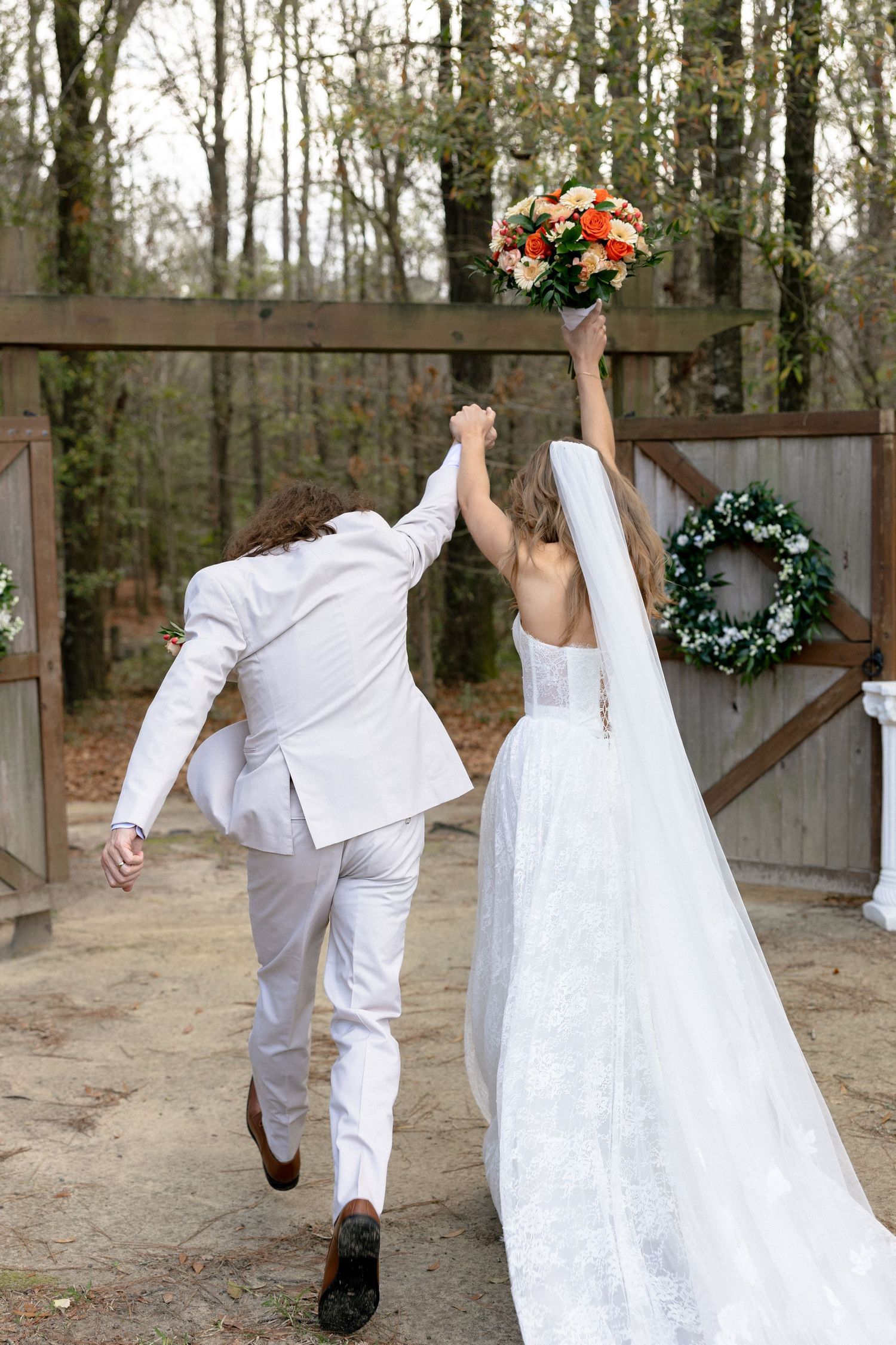 Newlyweds walk hand in hand through wooden gate, bride raising orange bouquet in celebration after ceremony.