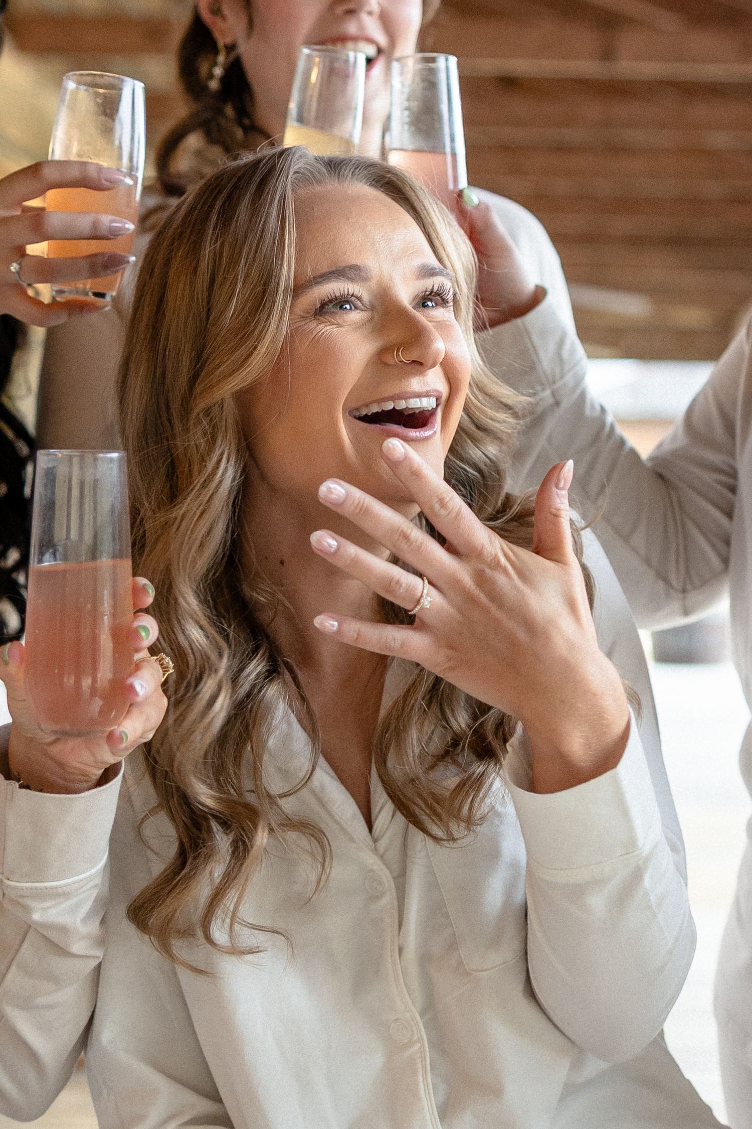 Excited bride showing engagement ring while holding champagne flute with bridesmaids.