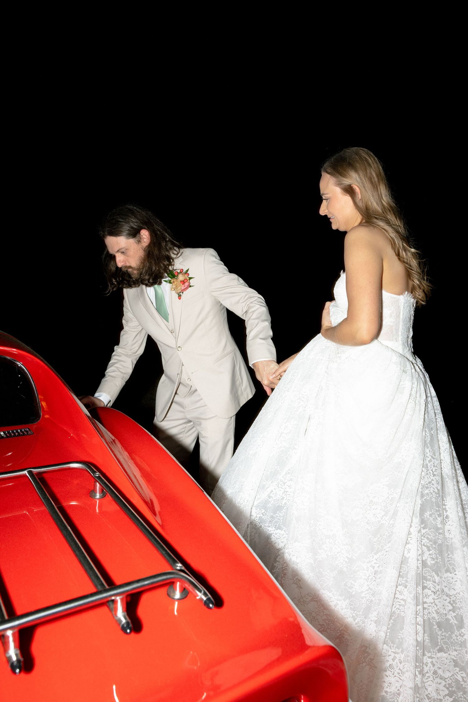 Newlyweds climbing into vintage red sports car after wedding ceremony at night.