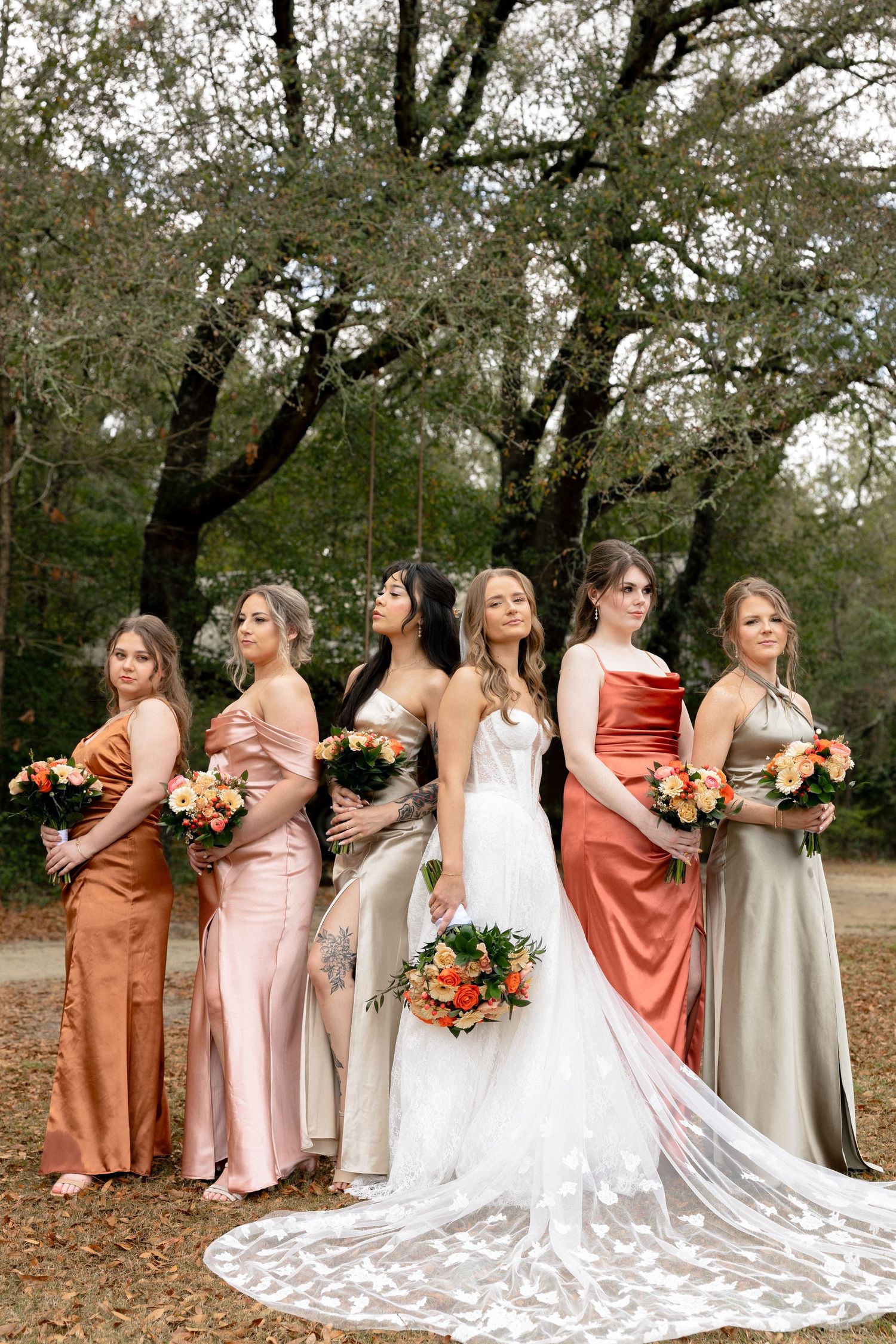 Bride and bridesmaids pose dramatically looking away in rust, blush, and sage satin gowns outdoors.