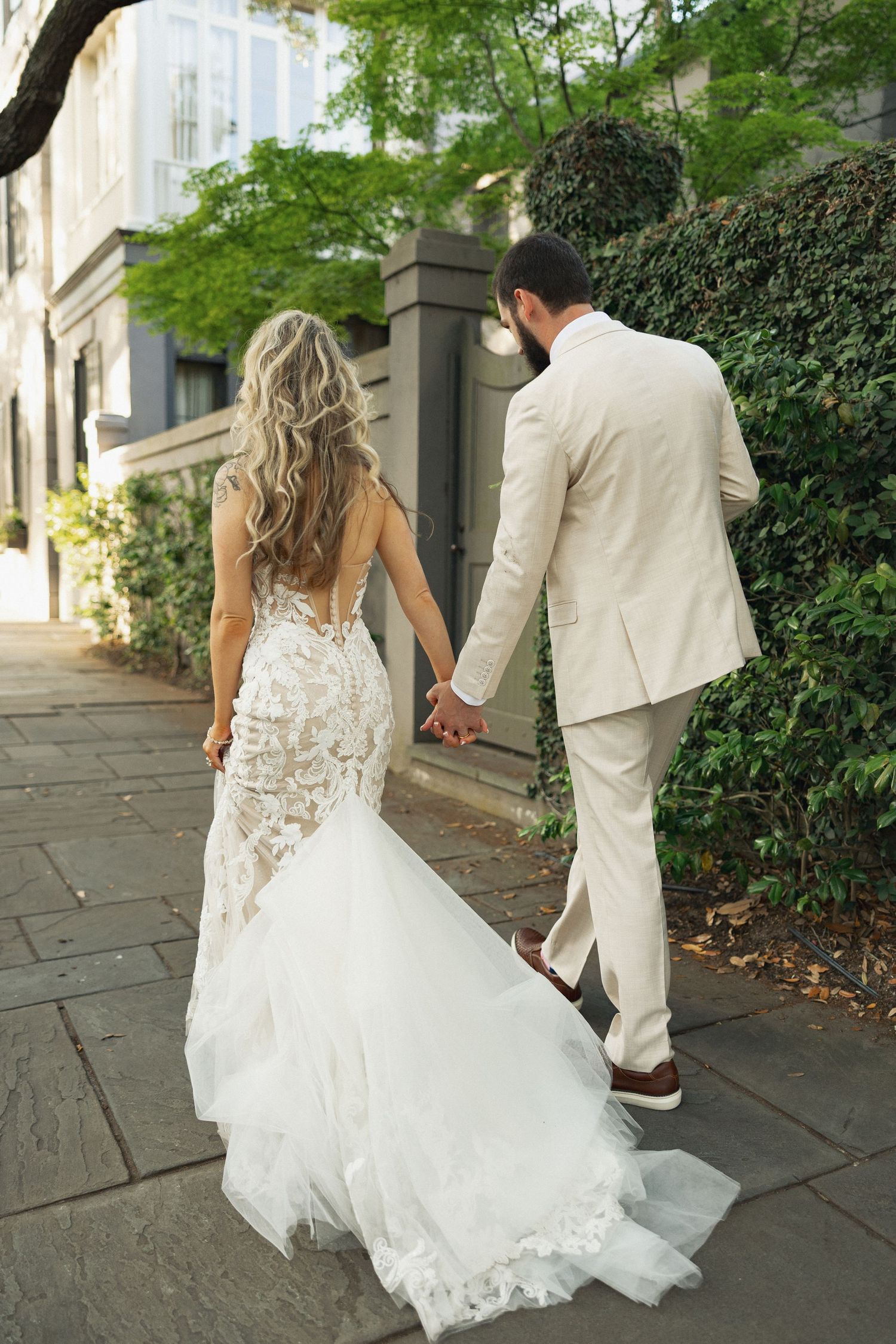 Bride and groom walk hand in hand on urban sidewalk, showing off lace backless wedding gown train.
