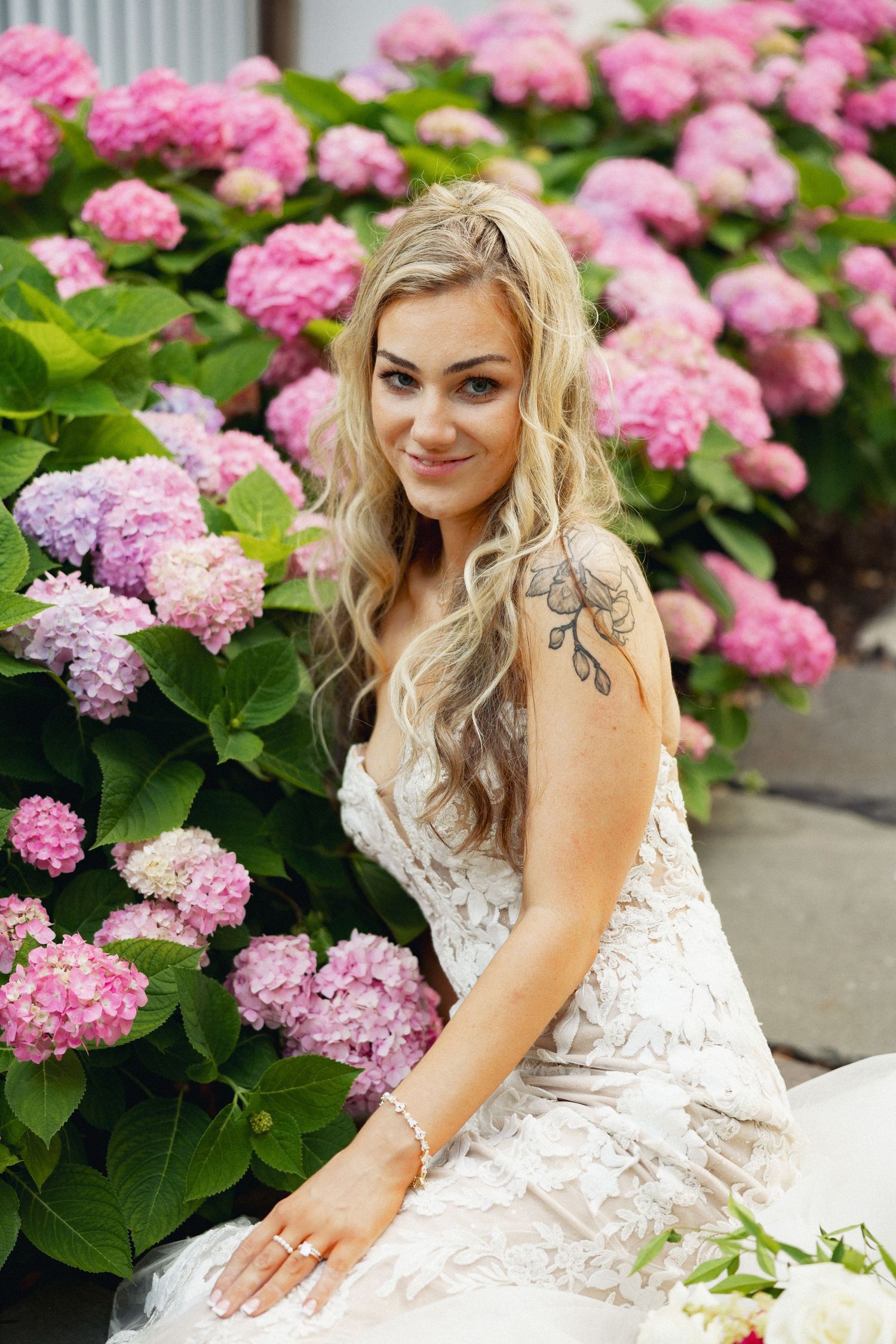 Smiling blonde bride in lace gown poses beside pink and purple hydrangea bushes outdoors.