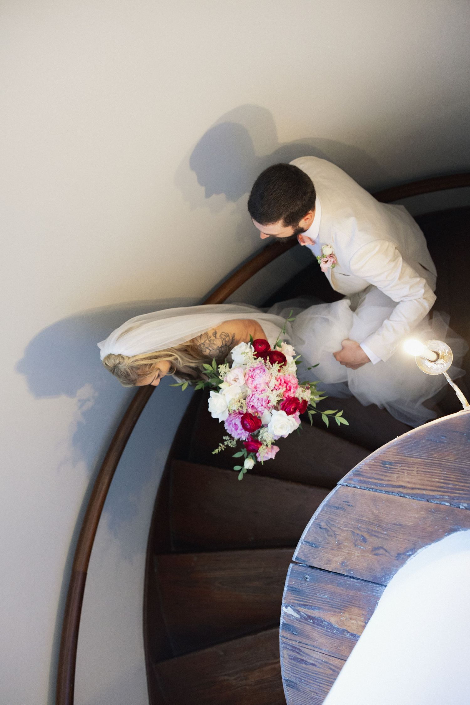 Bride and groom descend elegant spiral staircase, bride holding colorful wedding bouquet of pink and red flowers.