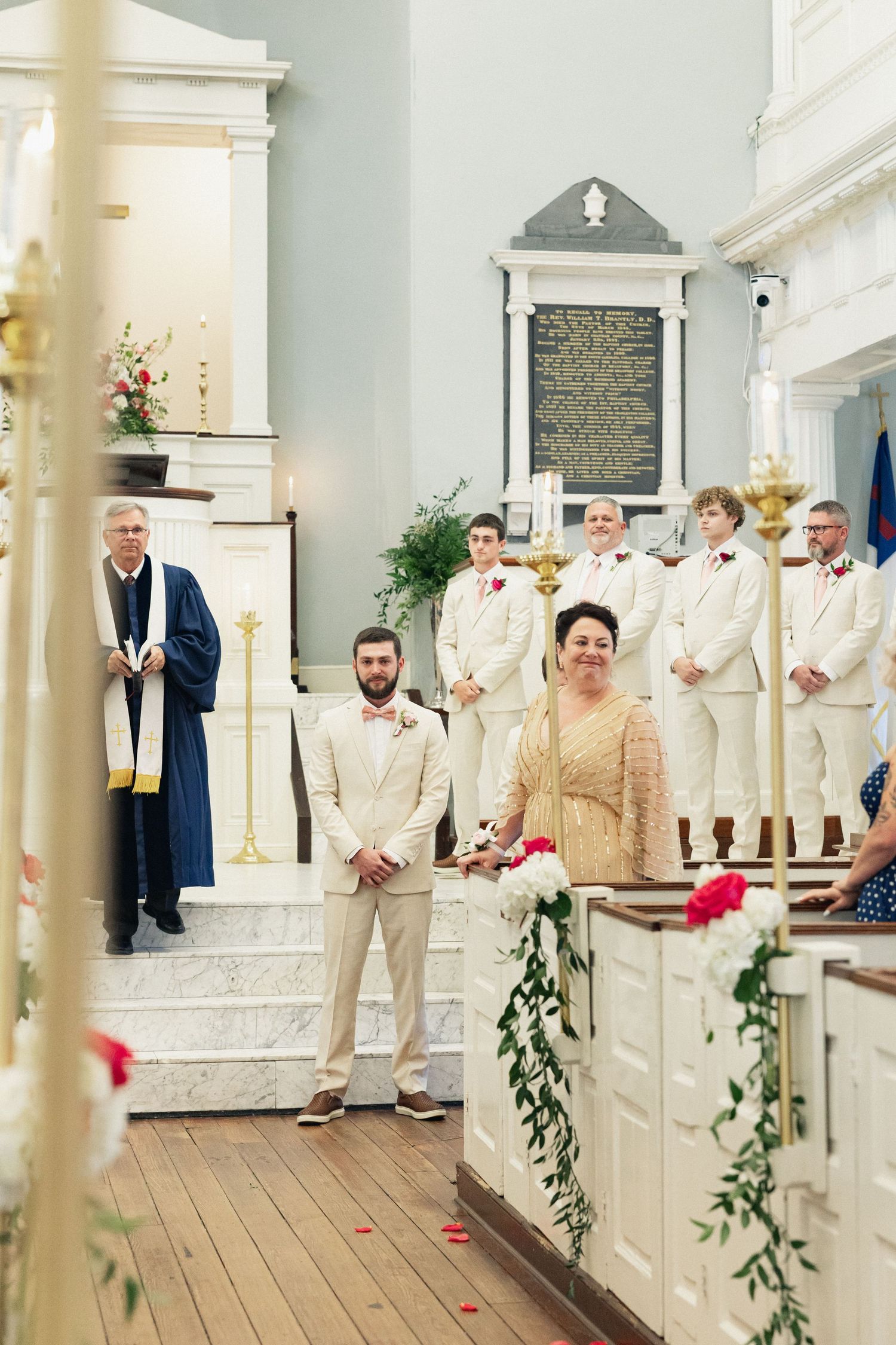 Groom in cream suit stands at church altar with groomsmen and officiant during traditional wedding ceremony.