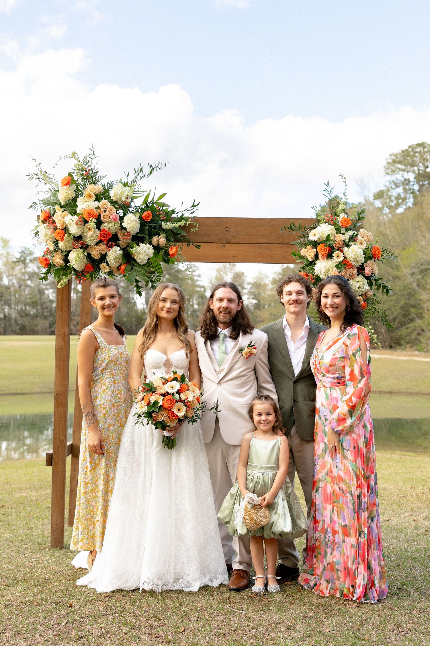 Wedding party poses under floral arch with orange and white blooms at outdoor ceremony.