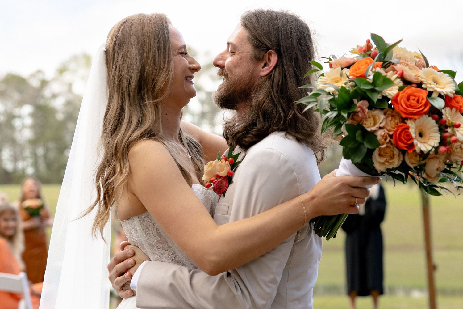 Smiling bride and groom embrace at outdoor wedding ceremony, bride holding vibrant orange and peach bouquet.