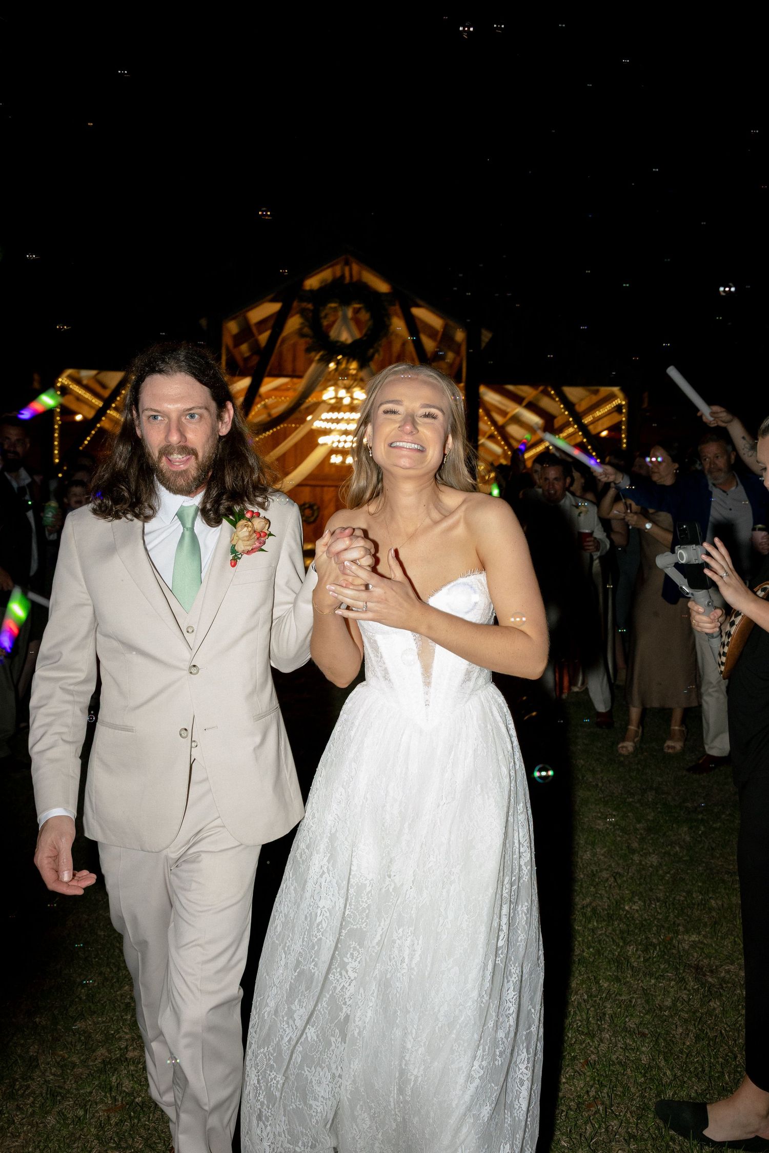 Newlyweds walking hand in hand during glowstick send-off at night wedding.