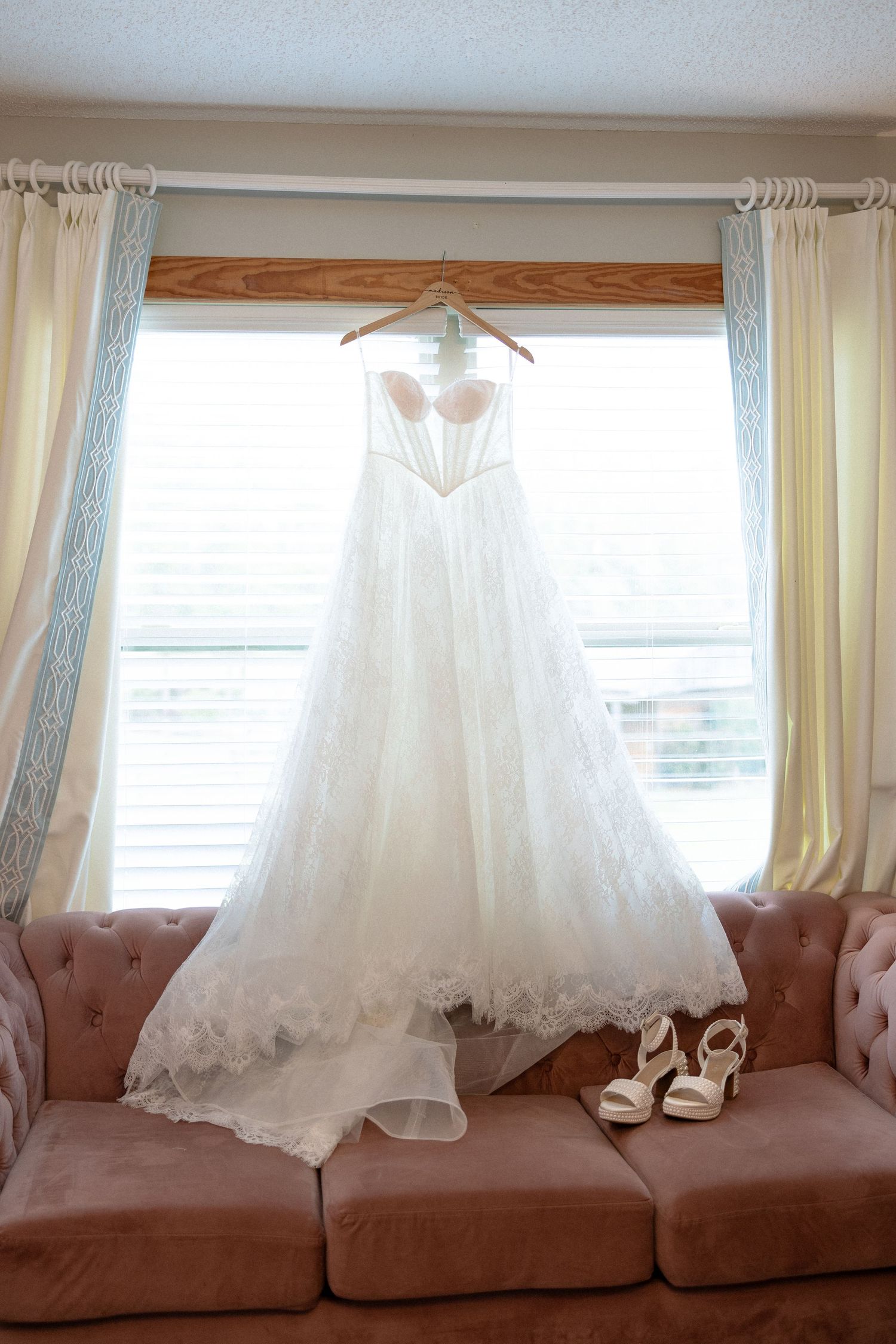 White strapless wedding gown hanging by a window above a pink sofa with bridal heels nearby.