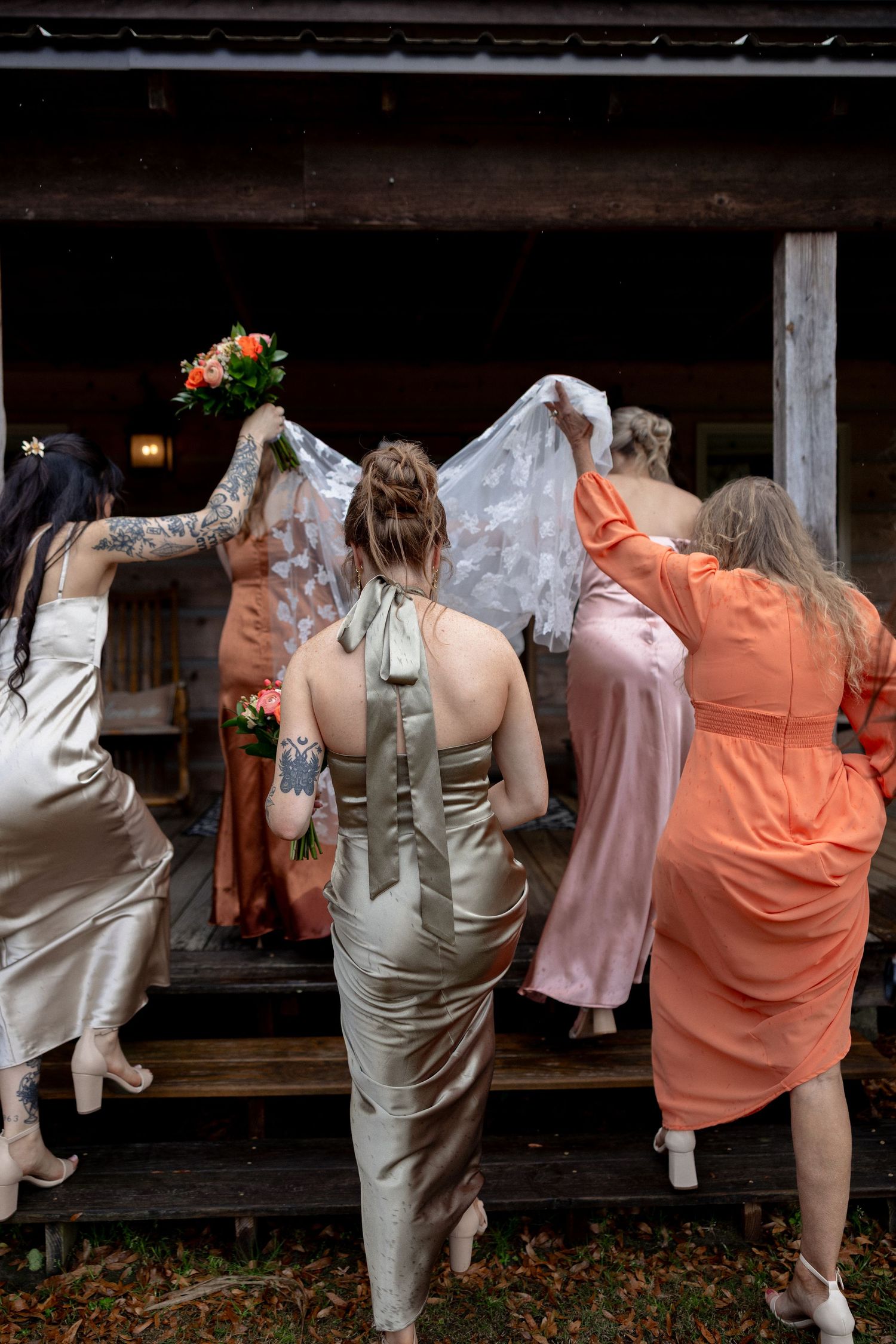 Bridesmaids in satin gowns help bride carry lace veil up wooden steps into dark rustic wedding venue.