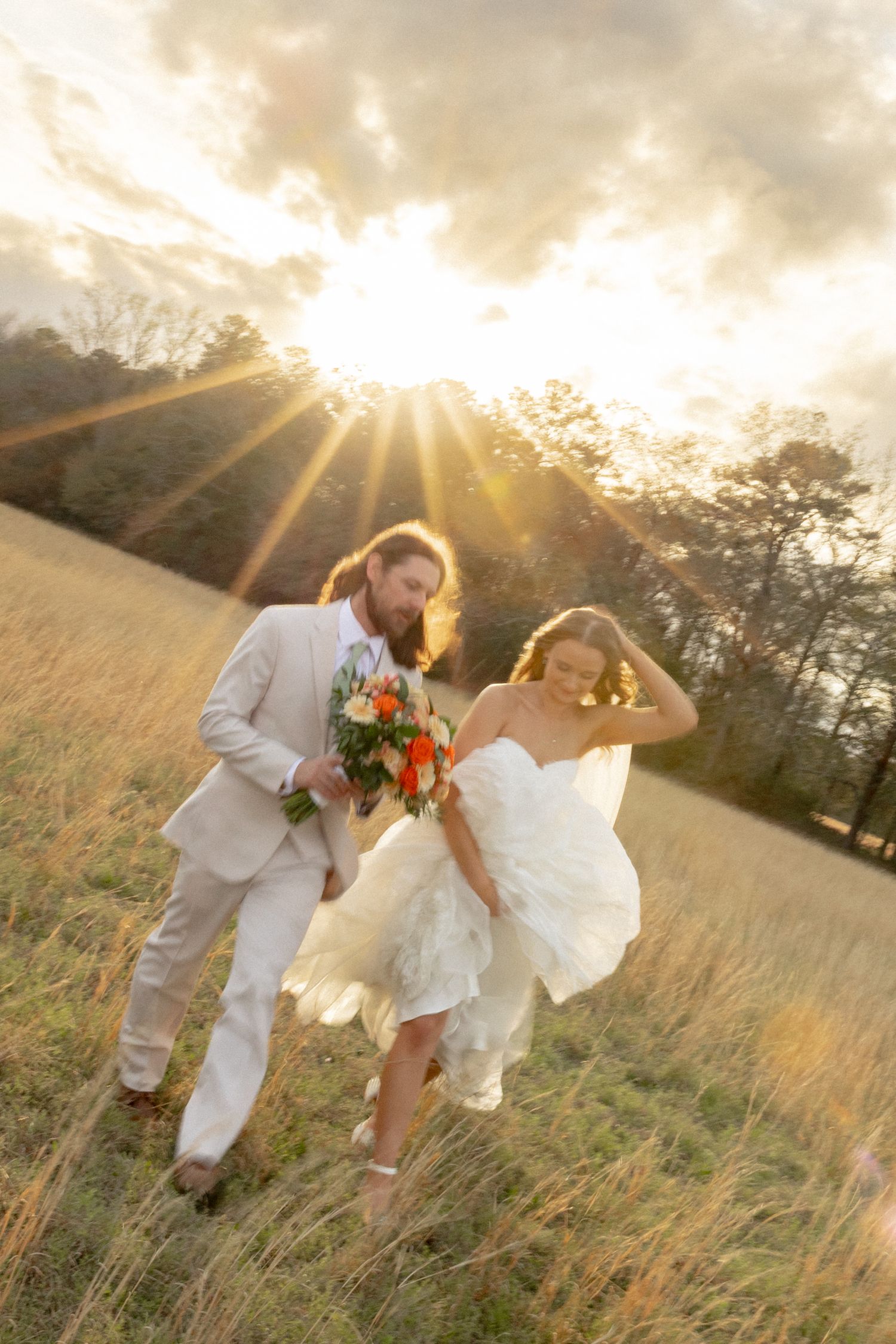 Joyful bride and groom running through a sunlit golden field at sunset, groom carrying colorful bridal bouquet.