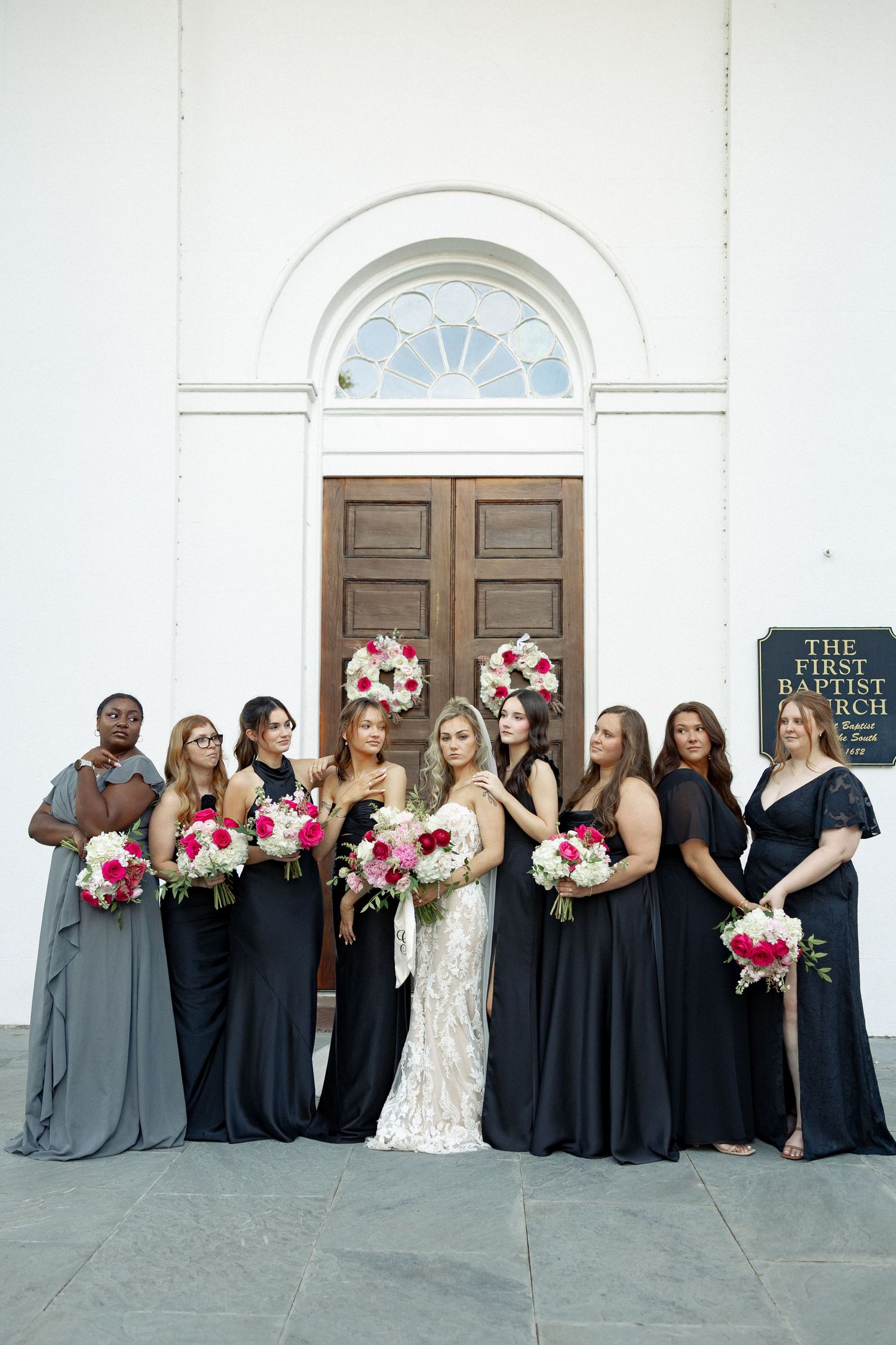 Bride in white lace gown with bridesmaids in black and gray dresses holding pink bouquets outside First Baptist Church.