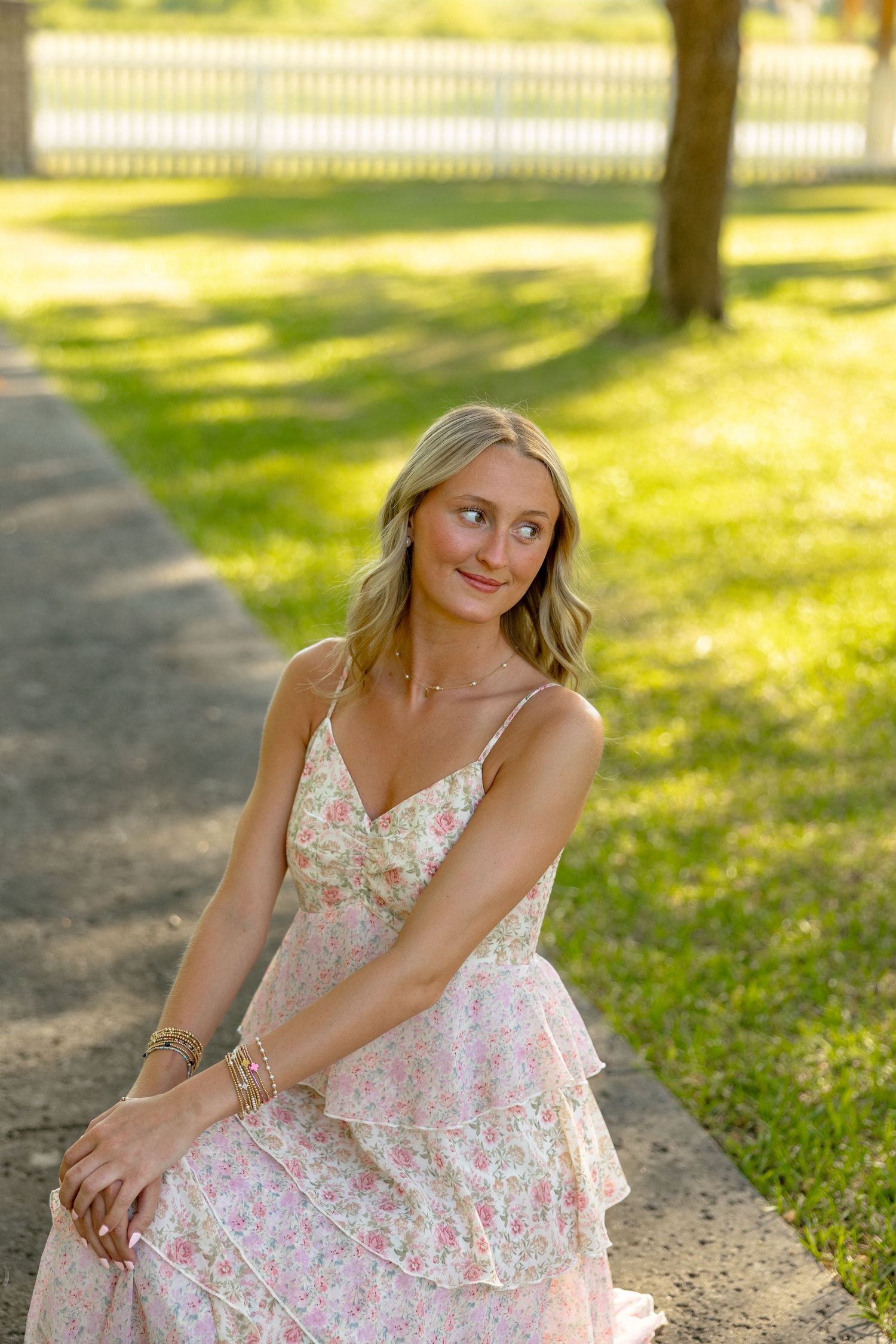 Young blonde woman in a pink floral dress sitting gracefully on a garden path in golden sunlight.