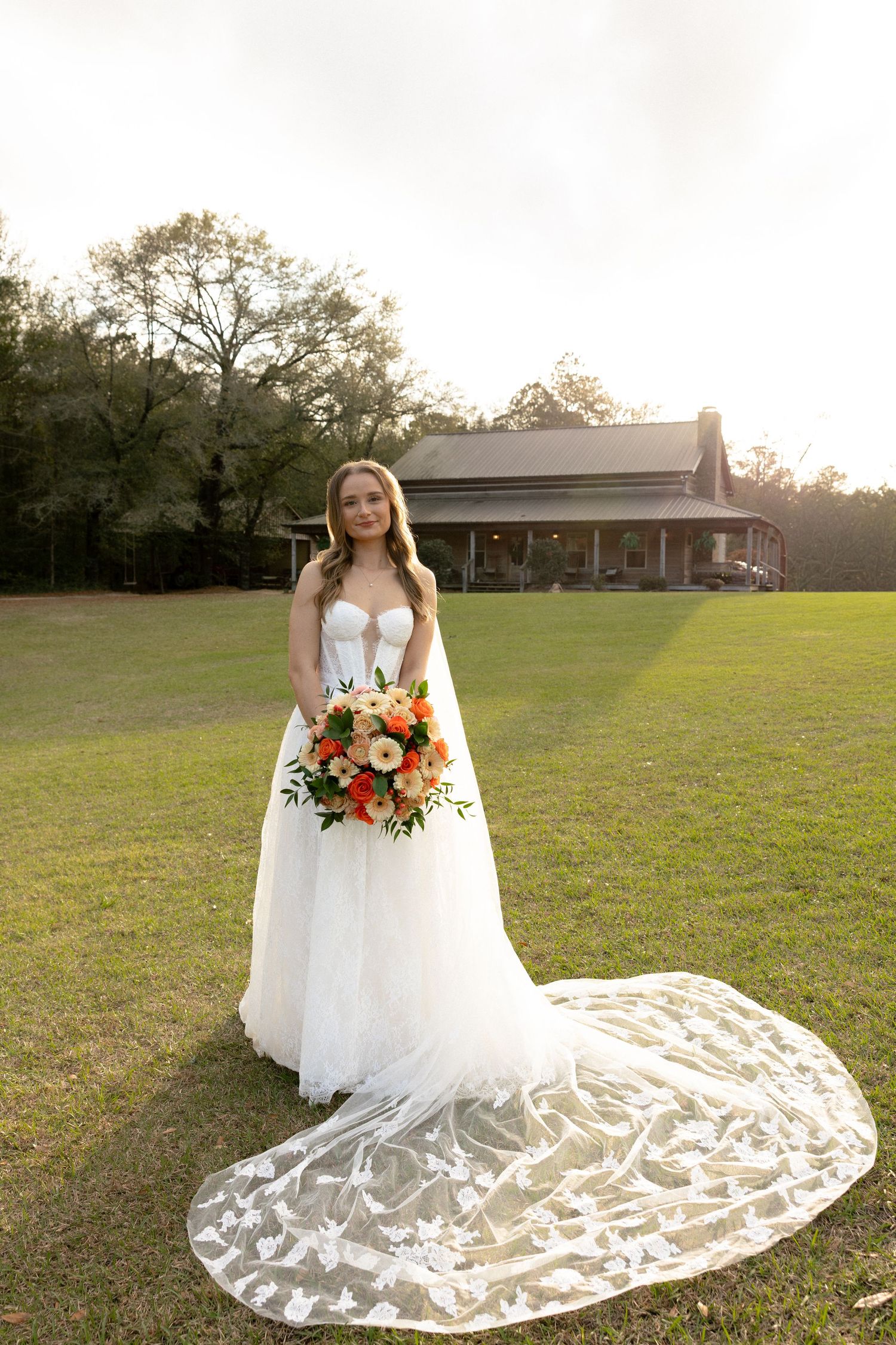 Bride in white lace gown with long train holding bouquet at golden hour.