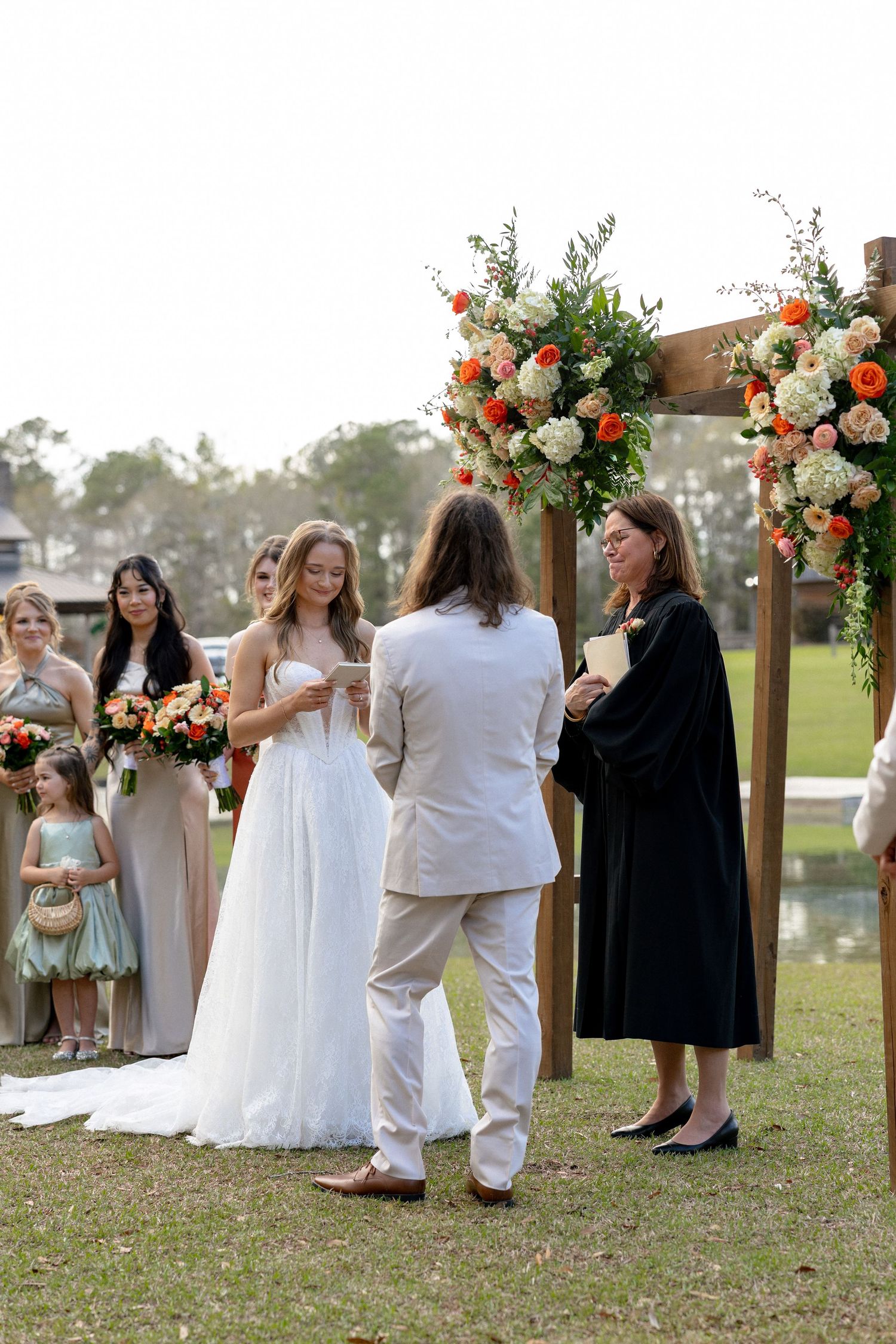 Bride reading vows to groom at outdoor wedding ceremony beneath wooden arch with orange floral arrangements.