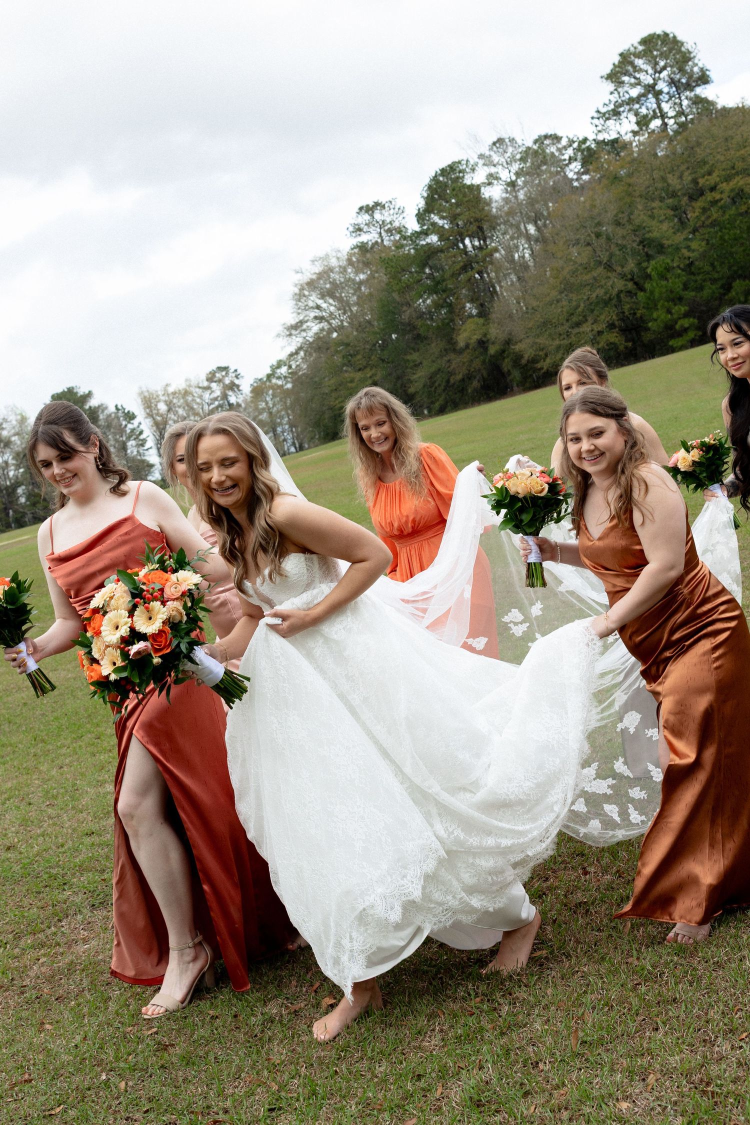 Bridesmaids in rust satin dresses help laughing bride lift her white lace gown while walking across green lawn.