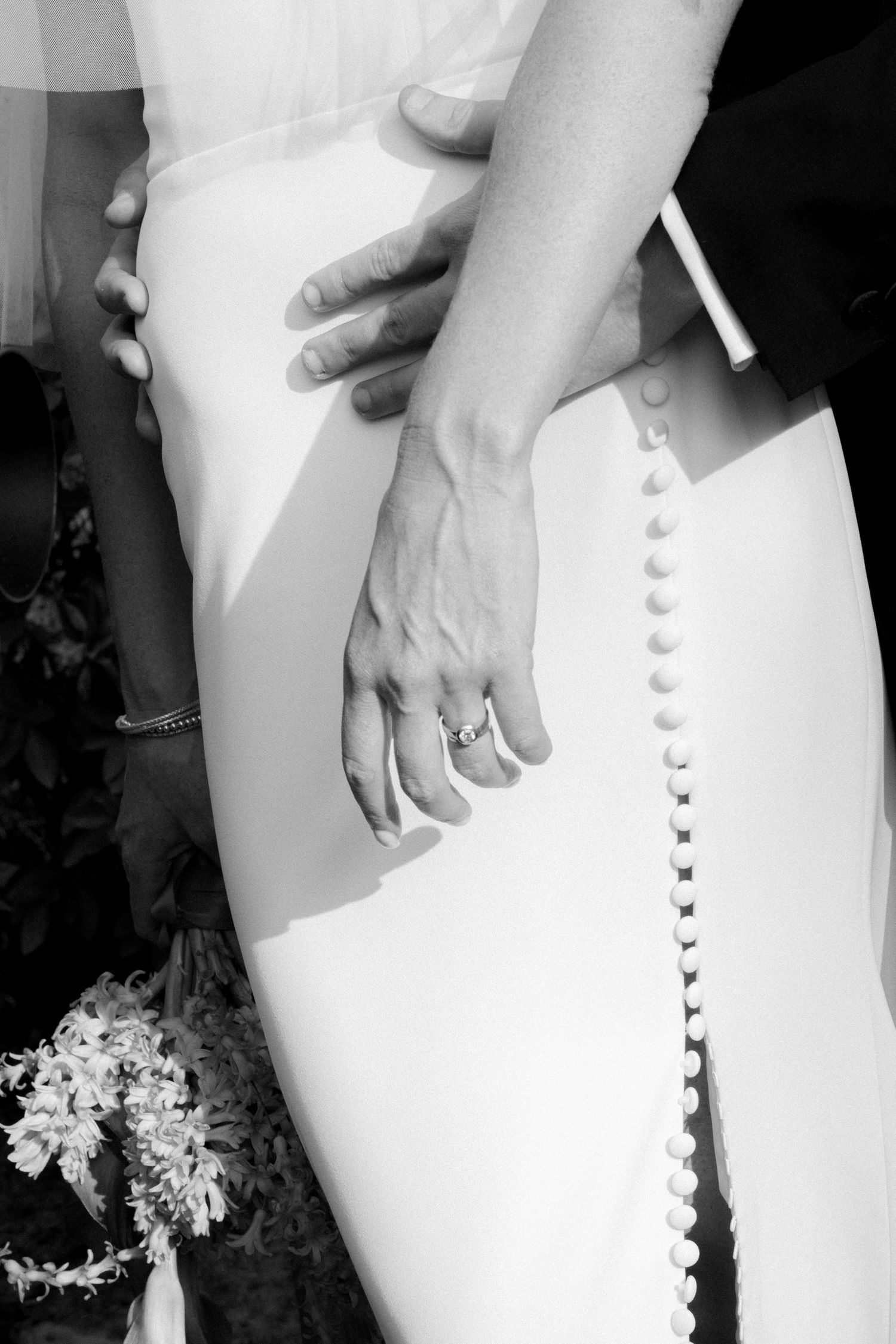 Black and white close-up of bride and groom hands, wedding ring visible on brides finger, button-back dress.