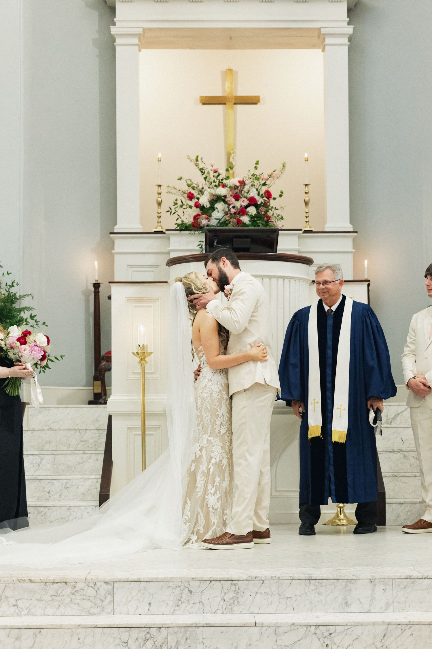Bride and groom share first kiss at elegant church wedding ceremony with pastor and cross altar.
