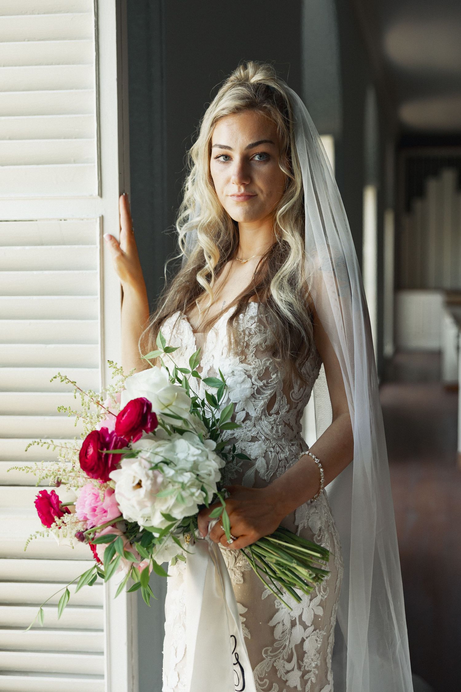 Blonde bride in lace wedding dress holding colorful bouquet of red and pink flowers by shuttered window.