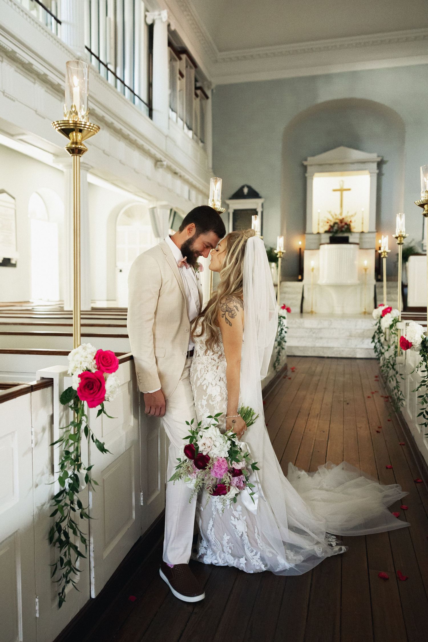 Bride and groom share a romantic moment in a historic white church, adorned with red floral arrangements.
