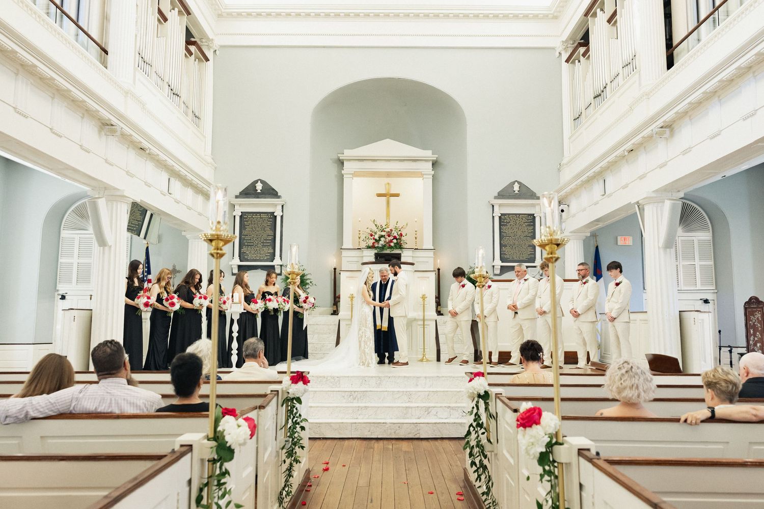 Full church view of wedding ceremony with bridal party at the altar, pews decorated with roses.