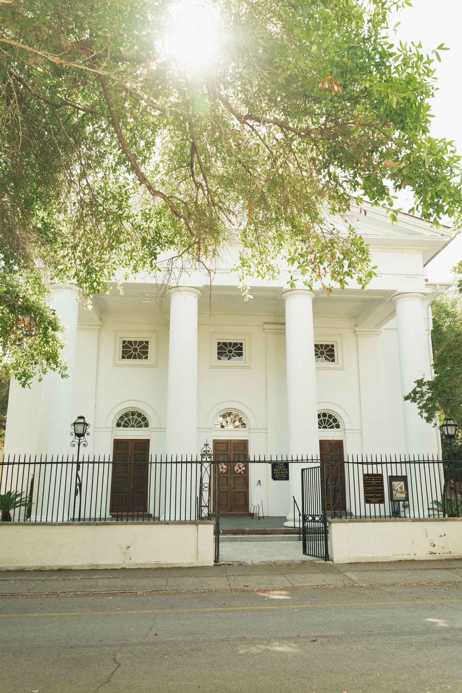 White neoclassical building with tall columns and arched windows, framed by lush green trees with sunlight filtering through.