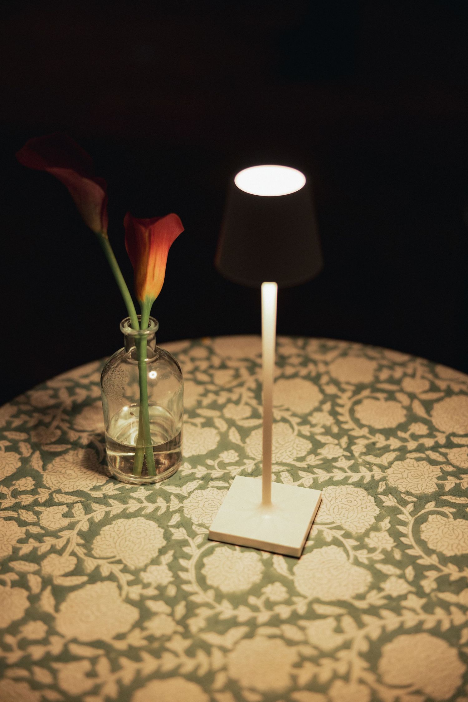 Small glowing table lamp beside a glass vase with red calla lilies on a green floral patterned tablecloth.