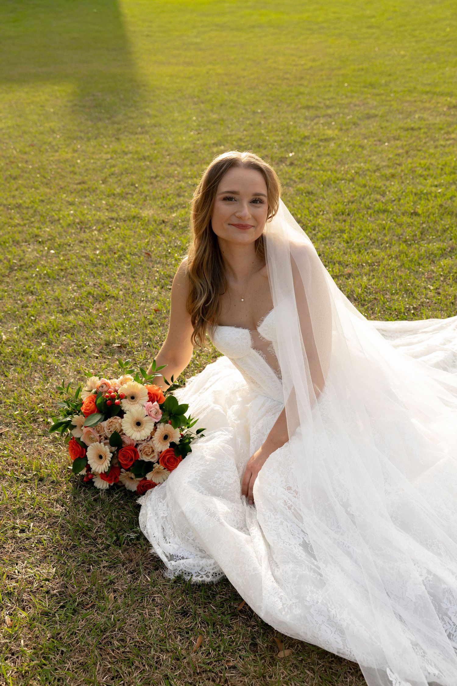 Bride seated on grass in white lace wedding dress holding vibrant bouquet.