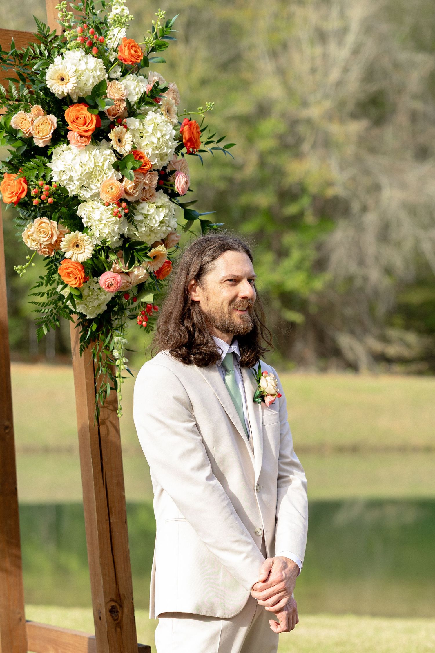 Groom in beige suit smiling at altar with orange and white floral arrangement at outdoor wedding ceremony.