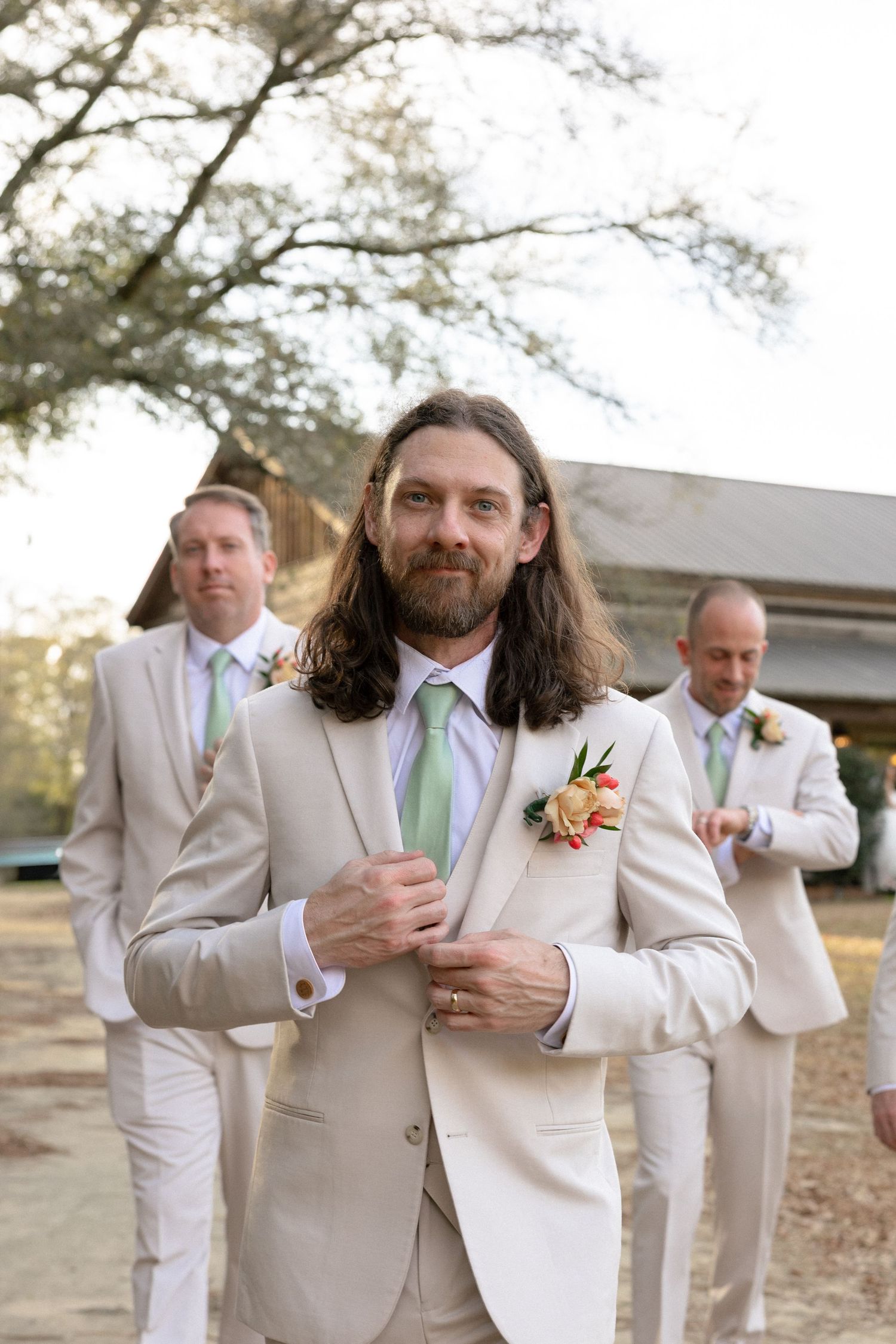 Groom in cream suit adjusts jacket while groomsmen stand behind him at a rustic barn wedding venue.