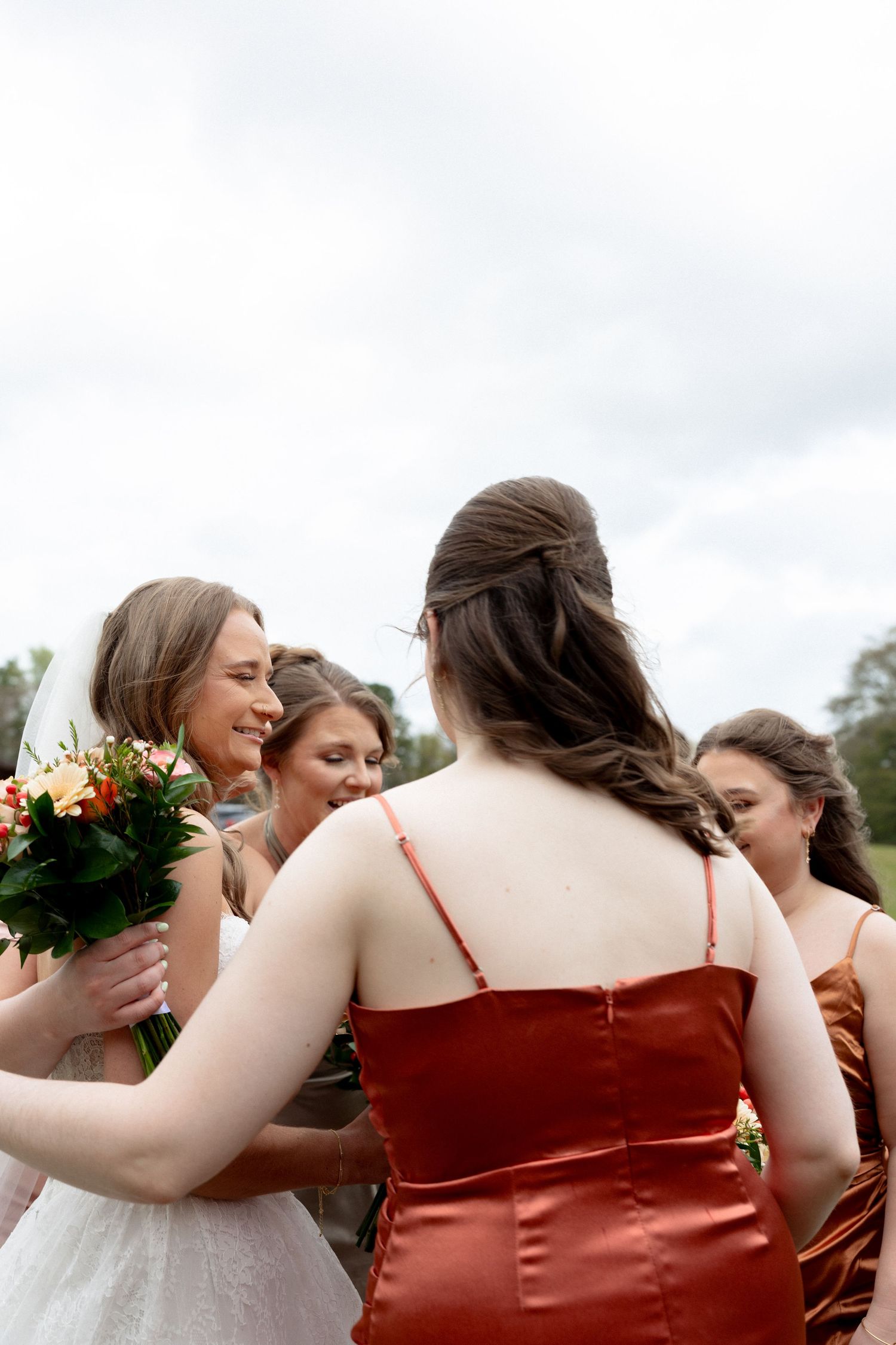 Bride and bridesmaids in rust and gold satin dresses share a joyful group moment outdoors at a wedding.