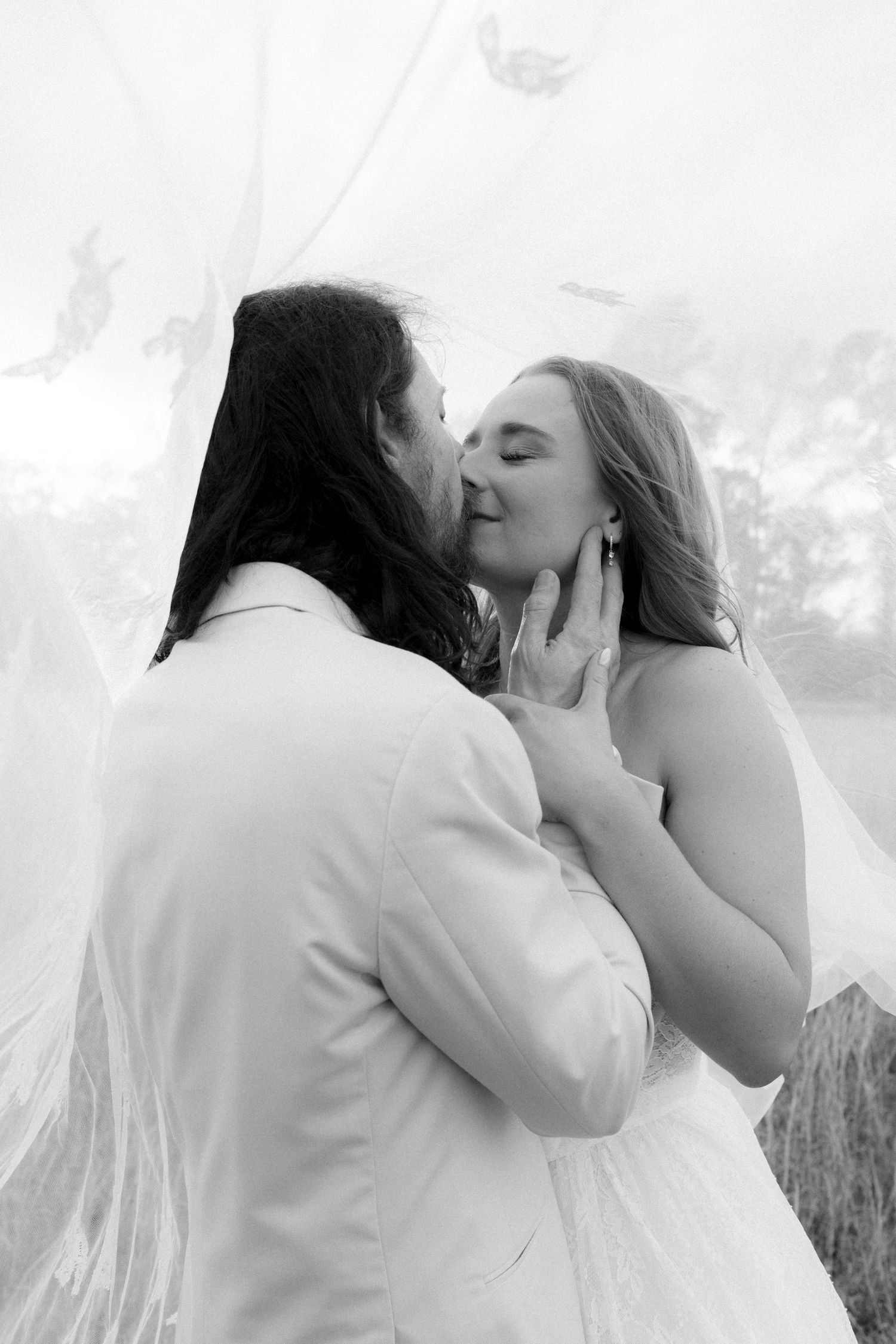 Black and white photo of groom tenderly kissing bride under flowing wedding veil.