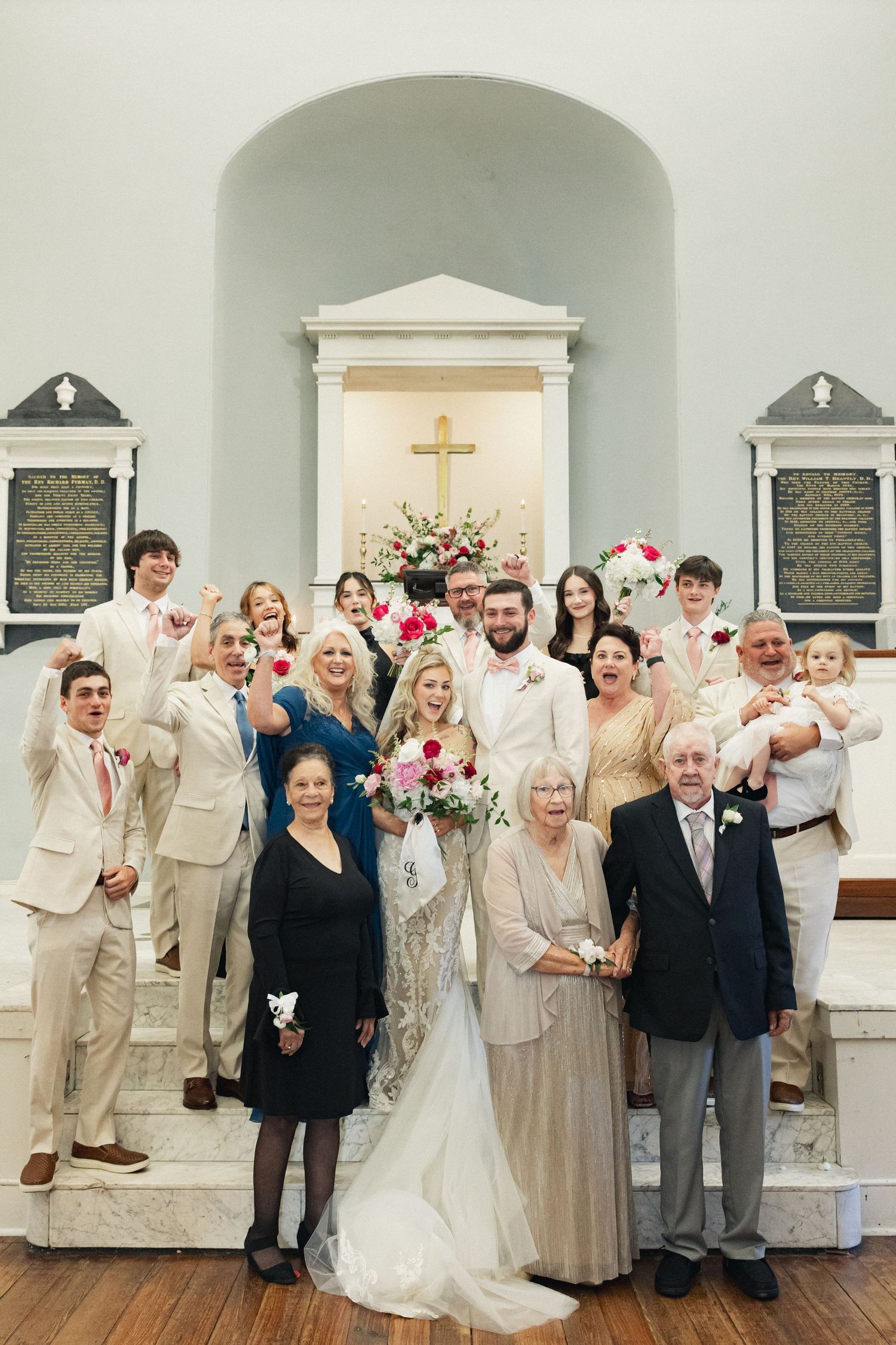 Wedding party poses with bride and groom before church altar with cross, family in formal attire celebrating joyfully.