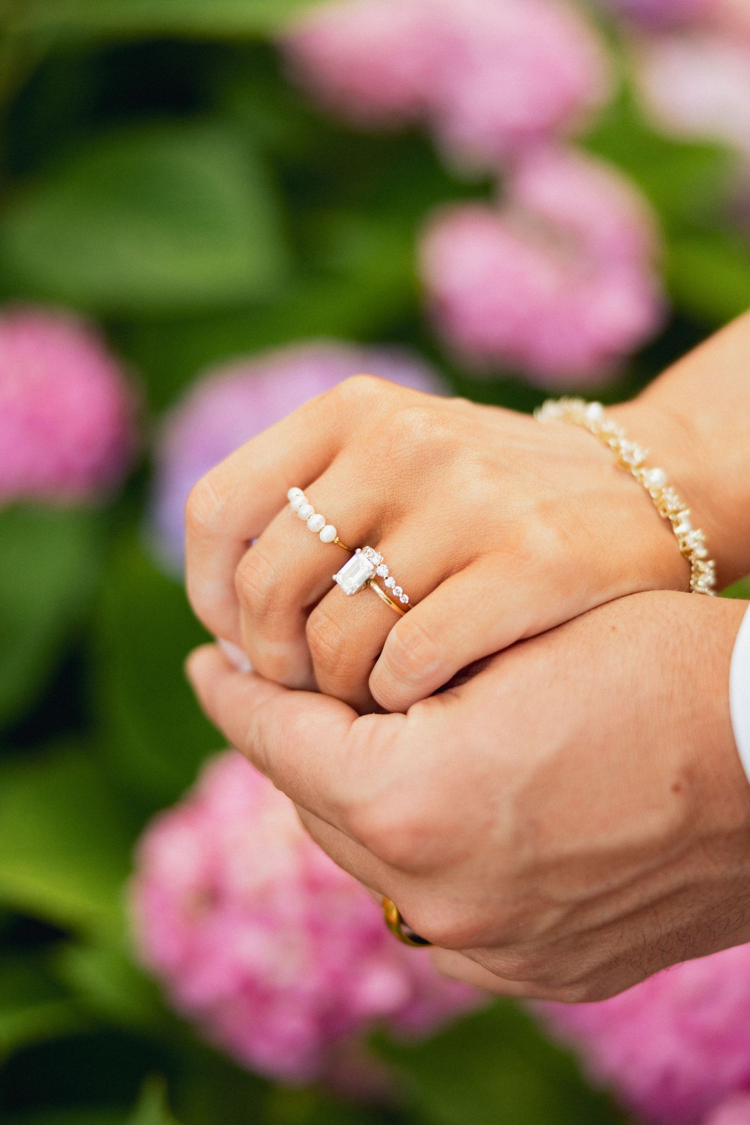 Close-up of bride's elegant emerald-cut diamond ring and pearl bracelet held by groom's hand near pink hydrangeas.