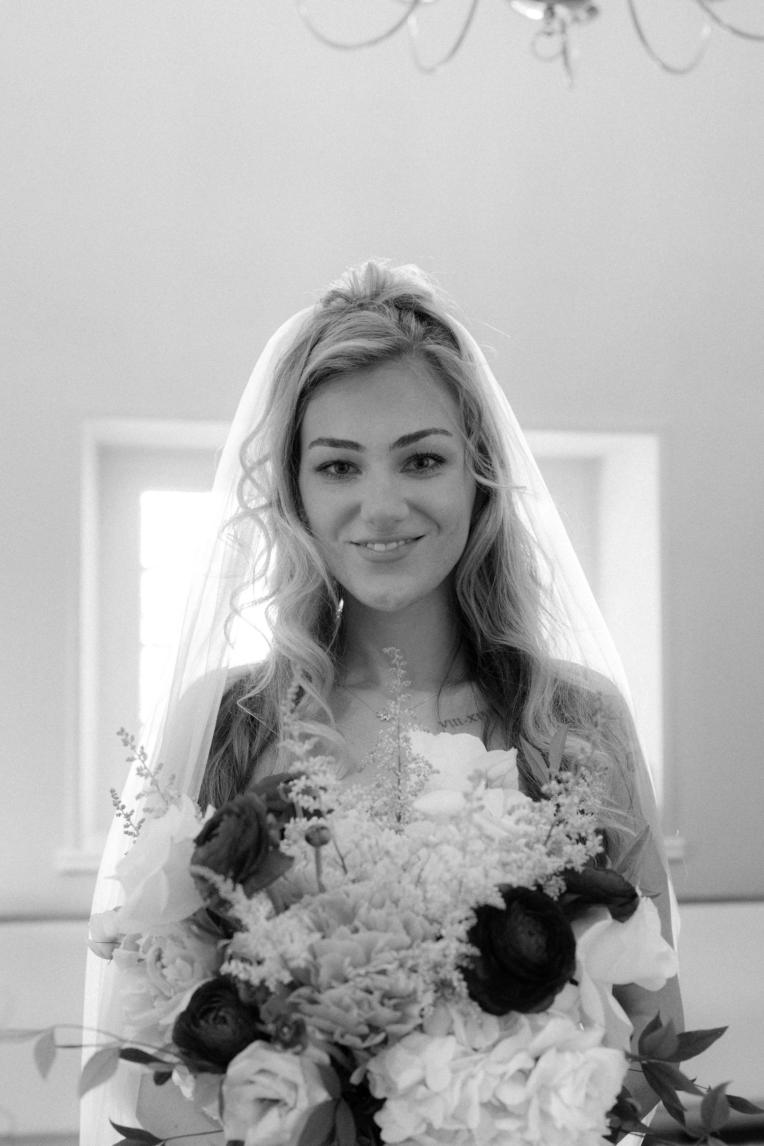 Black and white portrait of smiling bride in veil holding lush floral bouquet with roses.