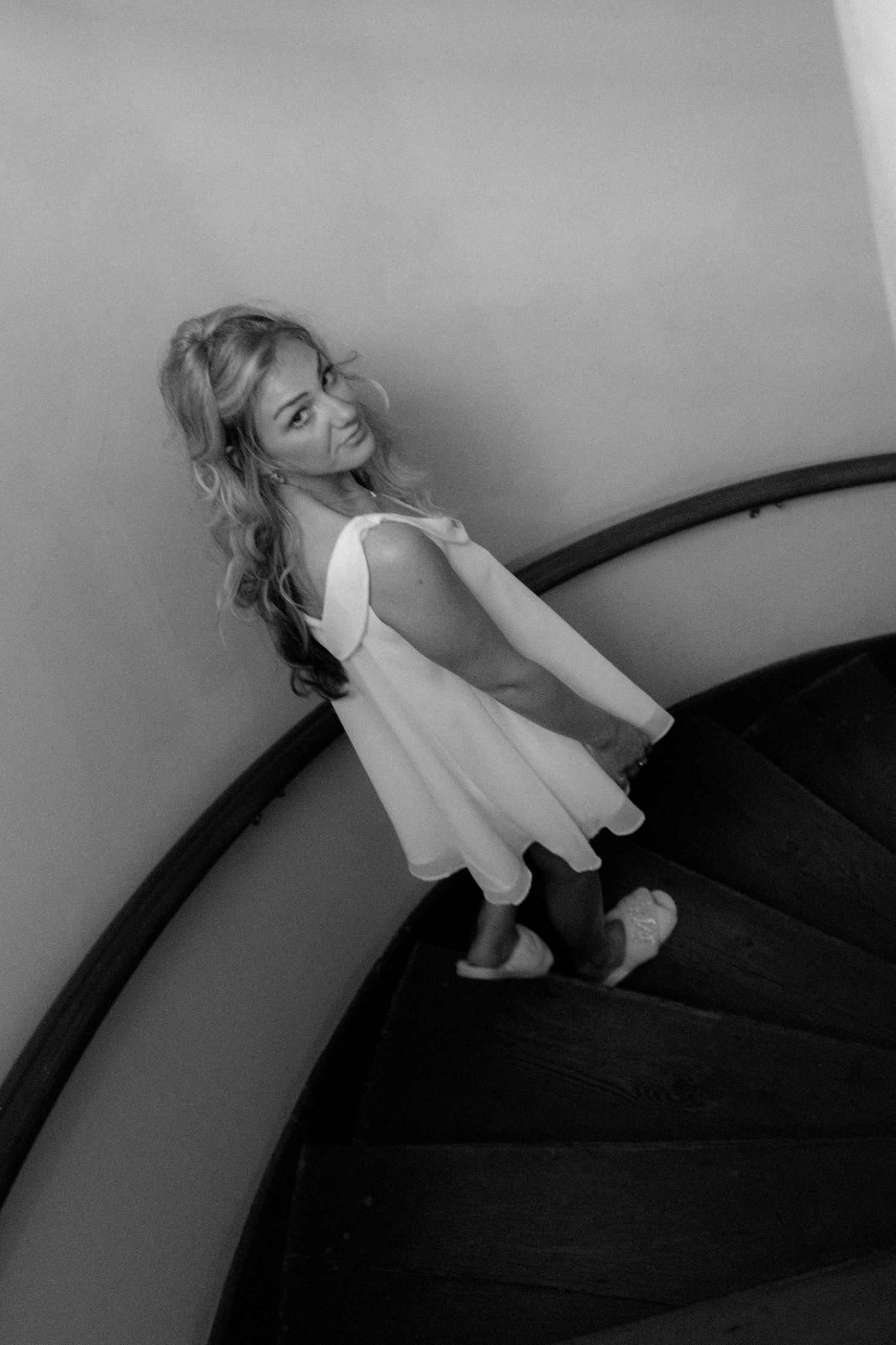 Black and white photo of a smiling young woman in a white dress on a spiral staircase.
