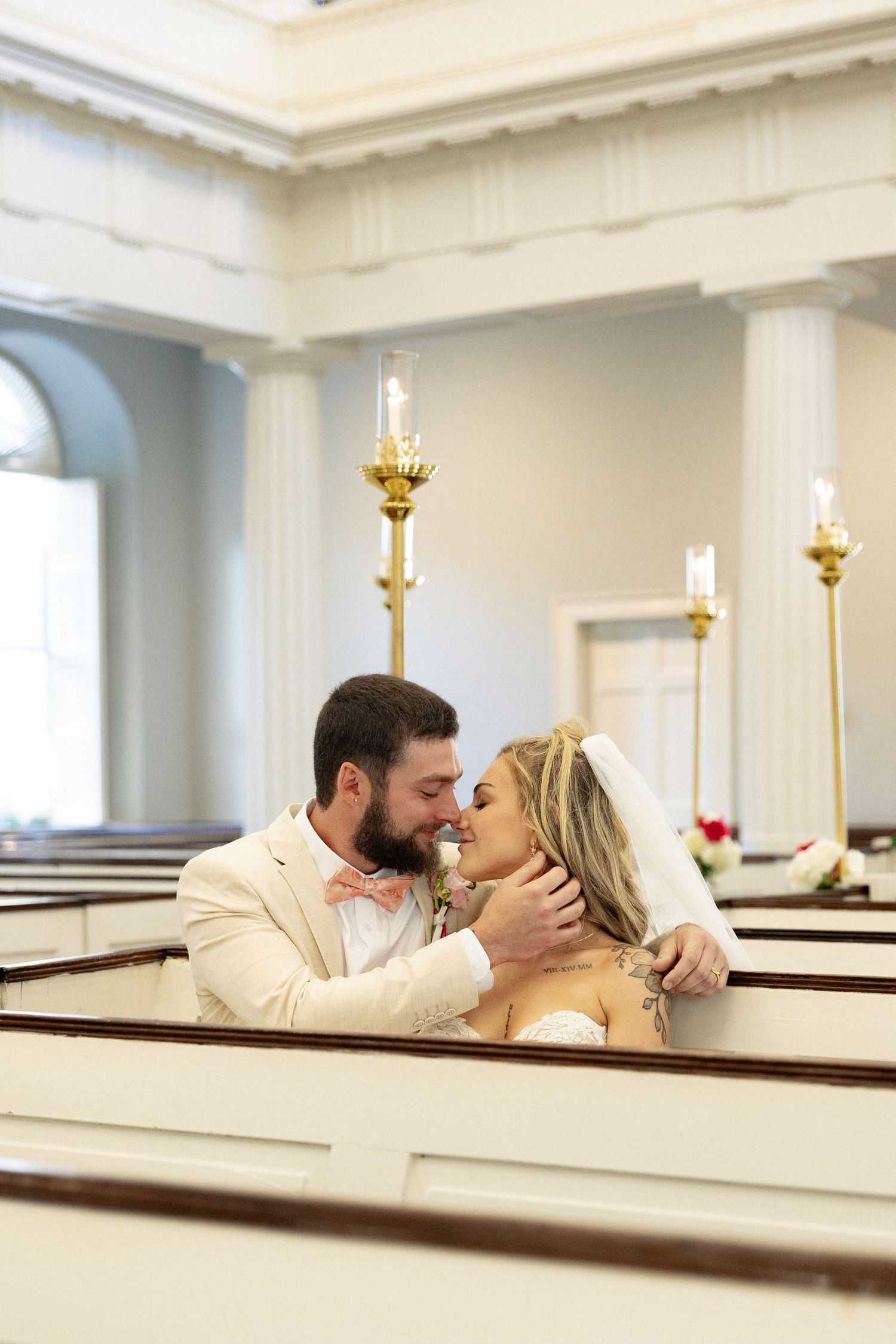 A bride and groom share a romantic moment in church pews, with the groom tenderly holding her face near golden candlesticks.
