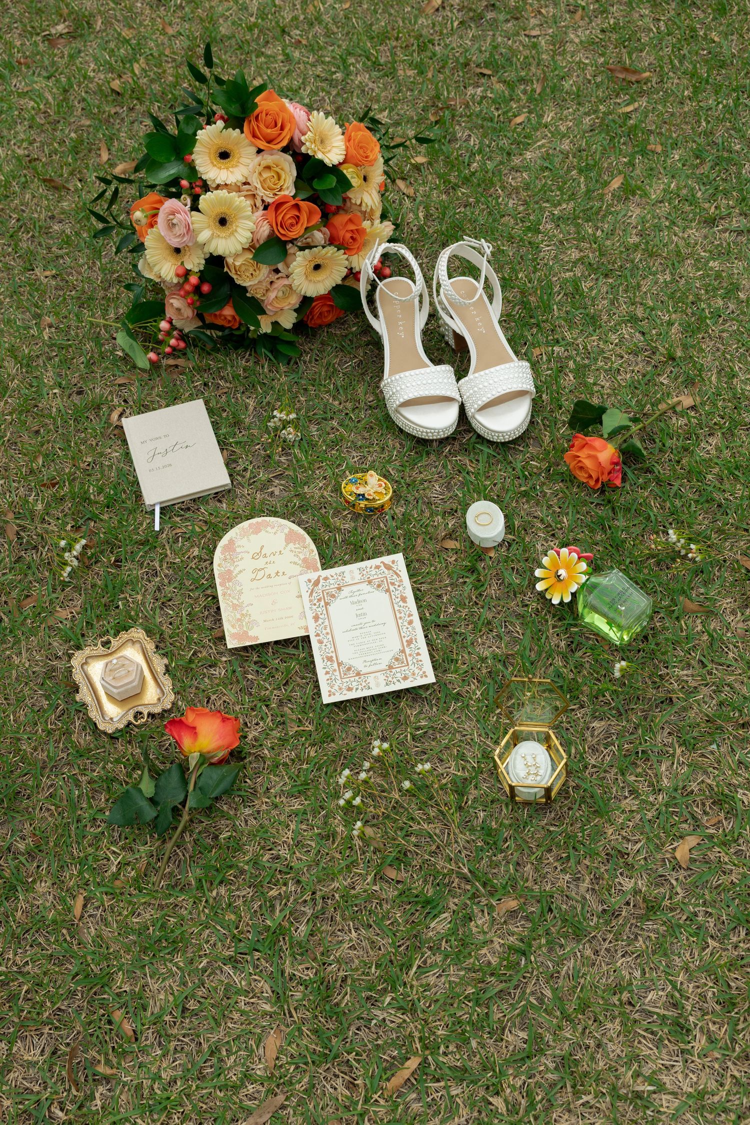 Bridal flat lay on green grass featuring orange bouquet, white heels, wedding invitations, rings, and accessories.