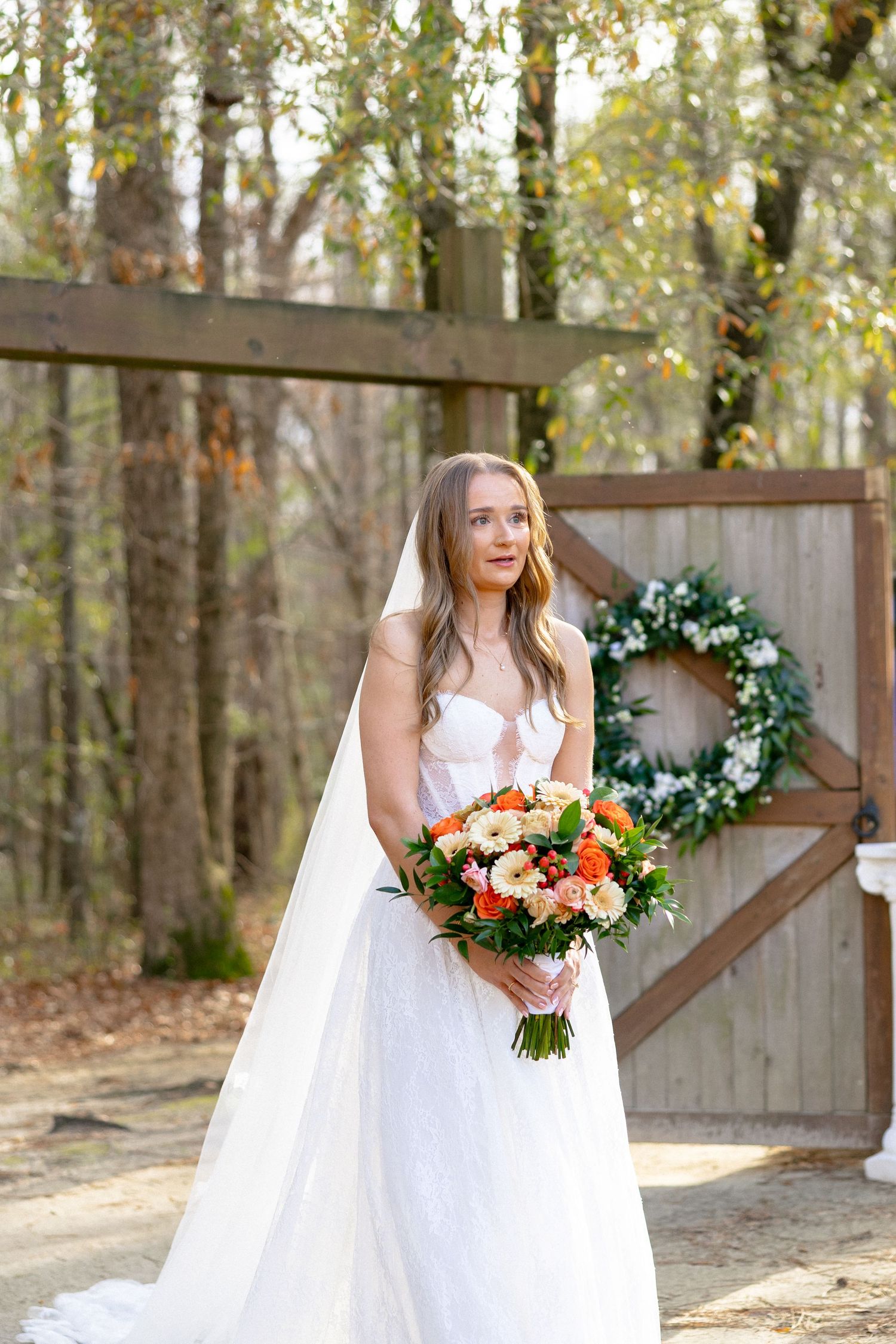 A beautiful bride in strapless white gown and veil holds colorful bouquet standing before a wooden gate with floral wreath.