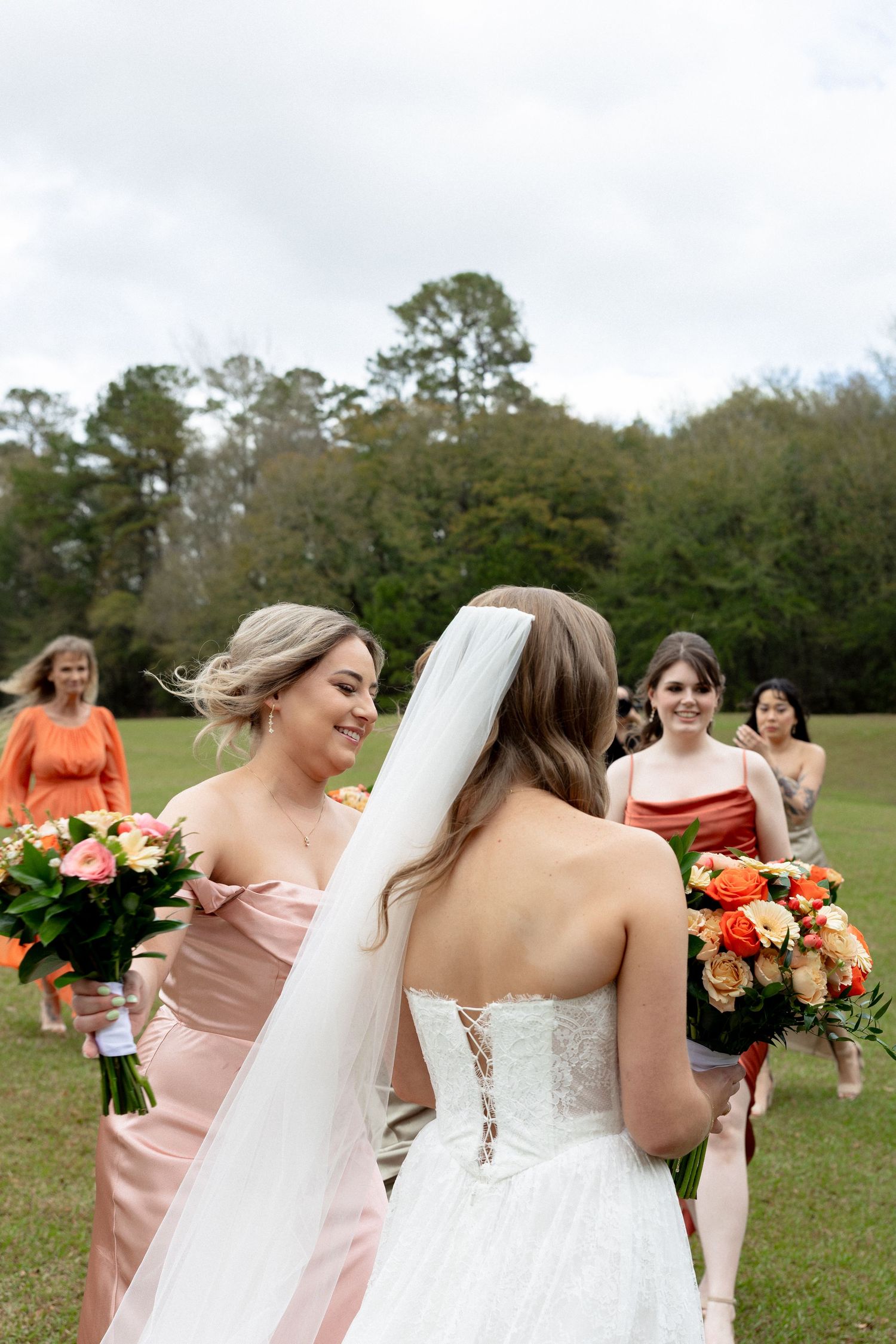 Bride in white lace gown with veil greets smiling bridesmaid in blush satin dress outdoors at wedding venue.