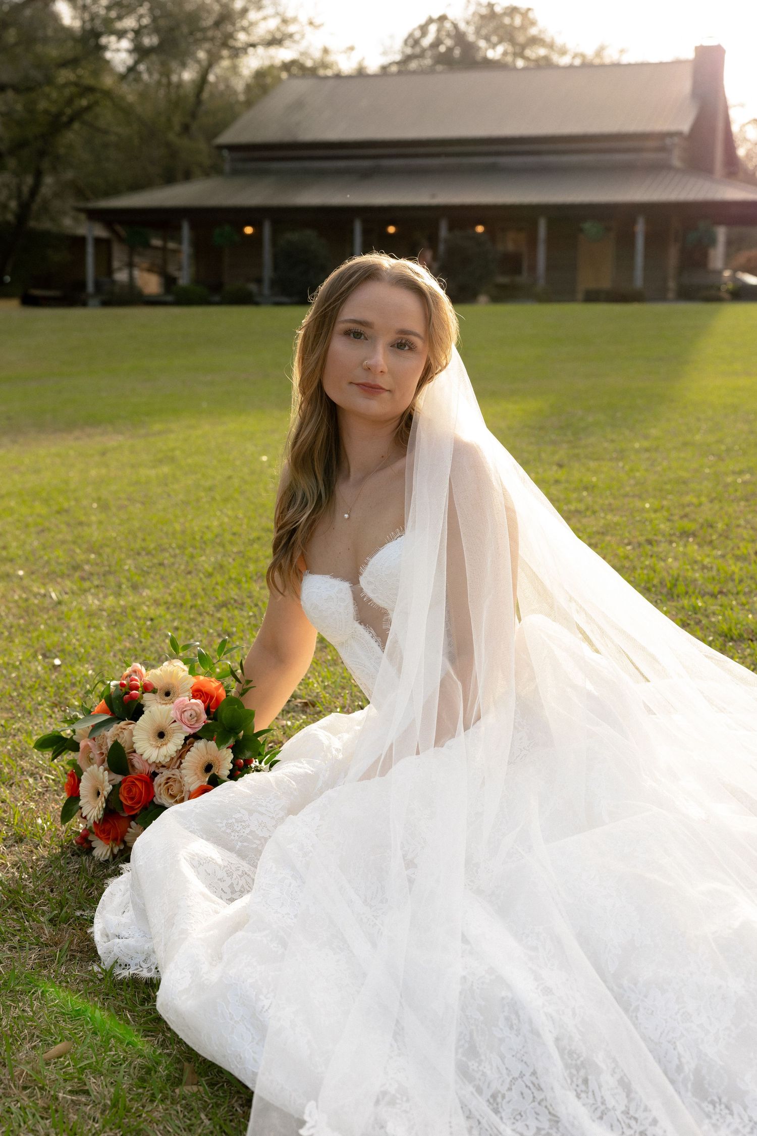 Bride in white lace gown with veil holding colorful bouquet on lawn.