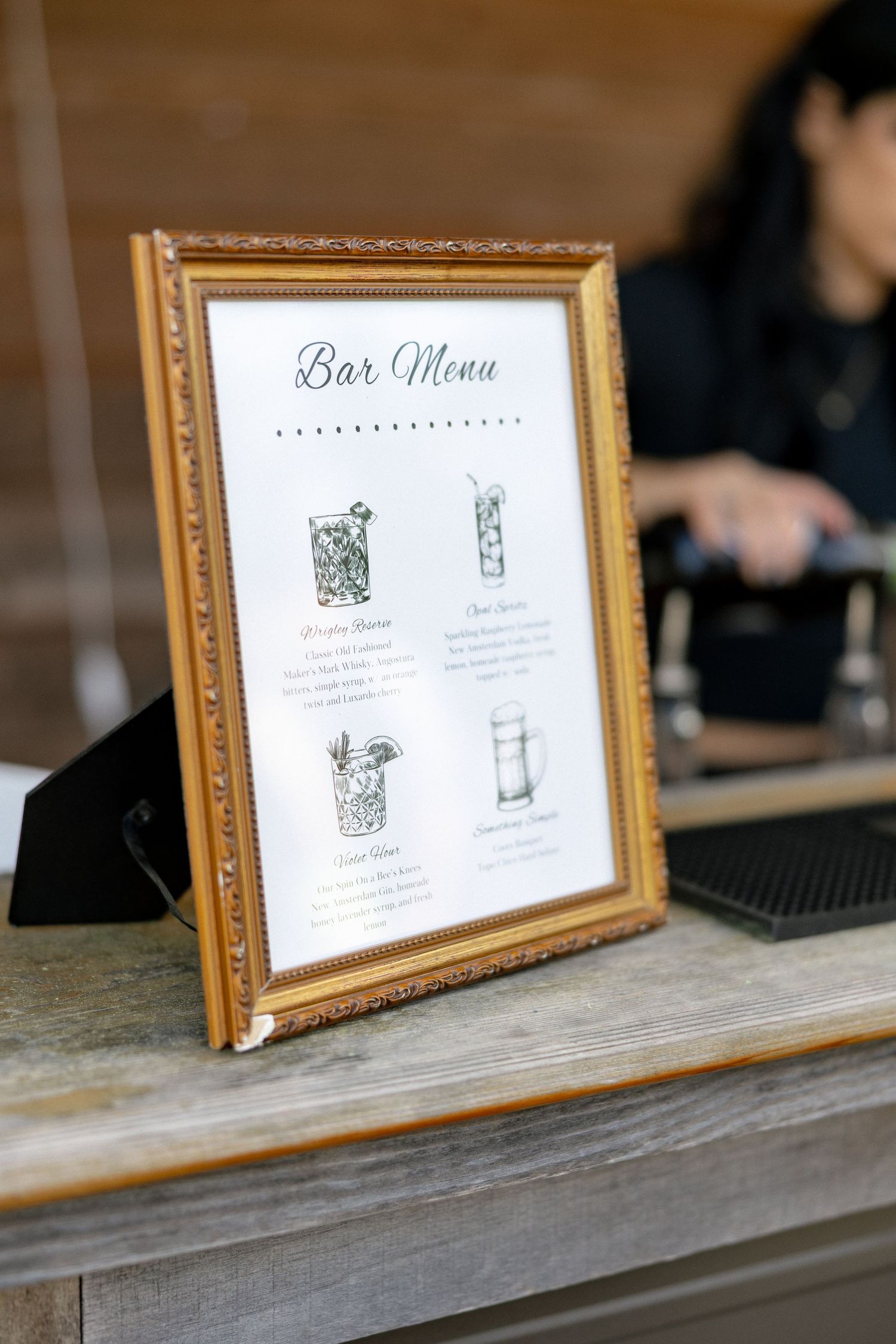 Elegant gold-framed bar menu sign displayed on a wooden counter at a wedding reception.