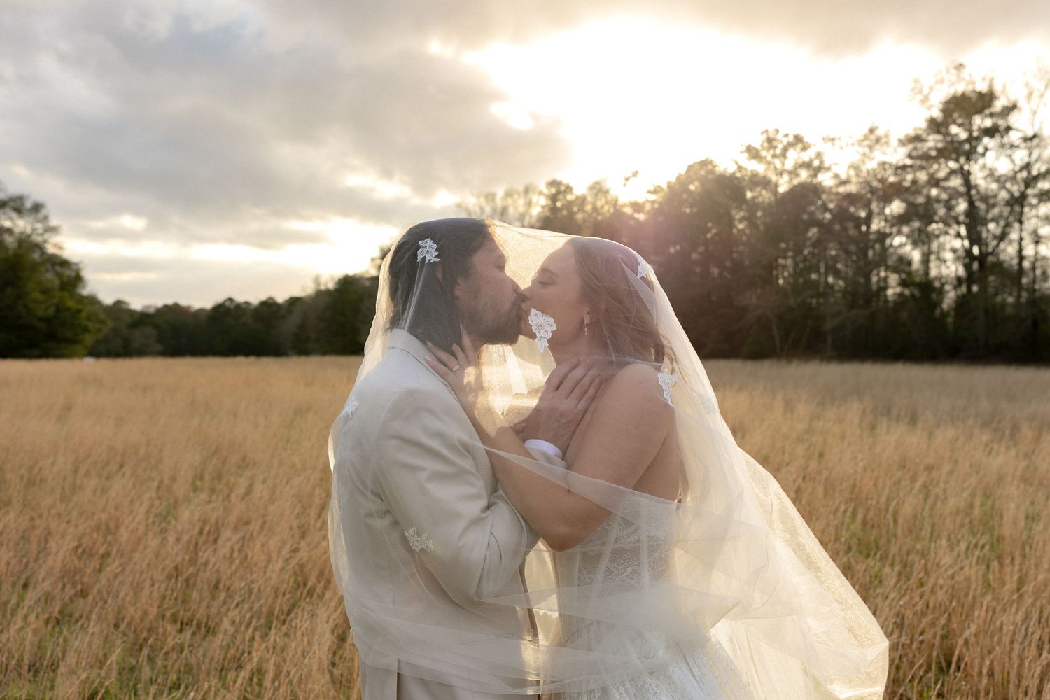 Bride and groom share a romantic moment under a veil in a golden field at sunset.