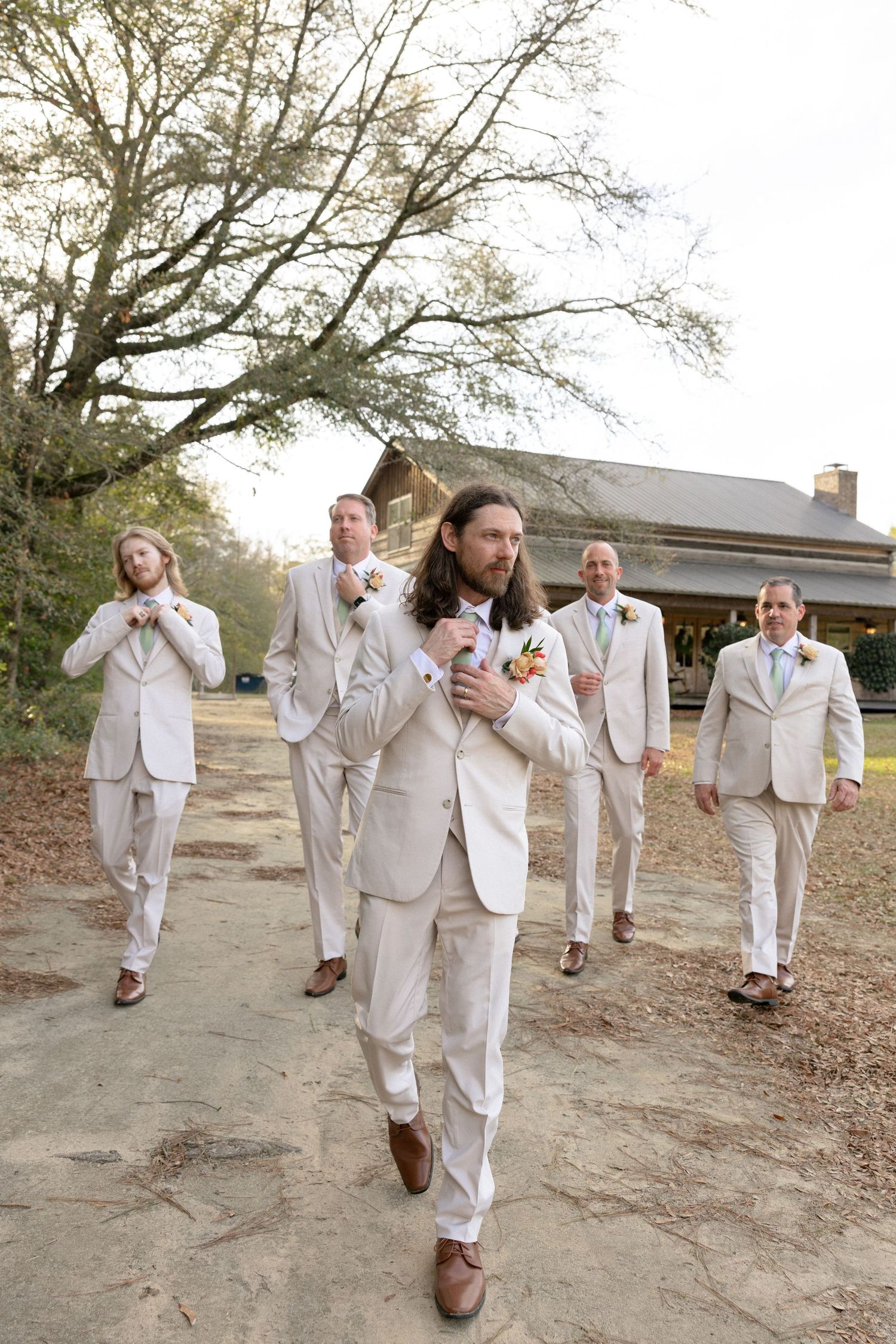 Groom in cream suit leads groomsmen walking down a path at a rustic outdoor barn wedding venue.