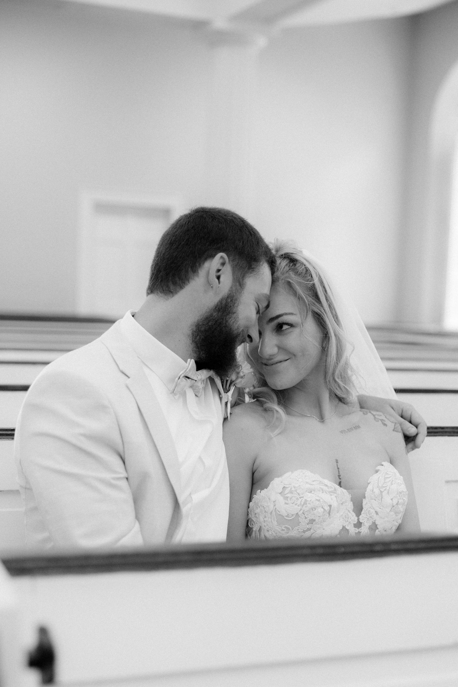 Black and white photo of a bearded groom nuzzling his smiling bride in a church pew.