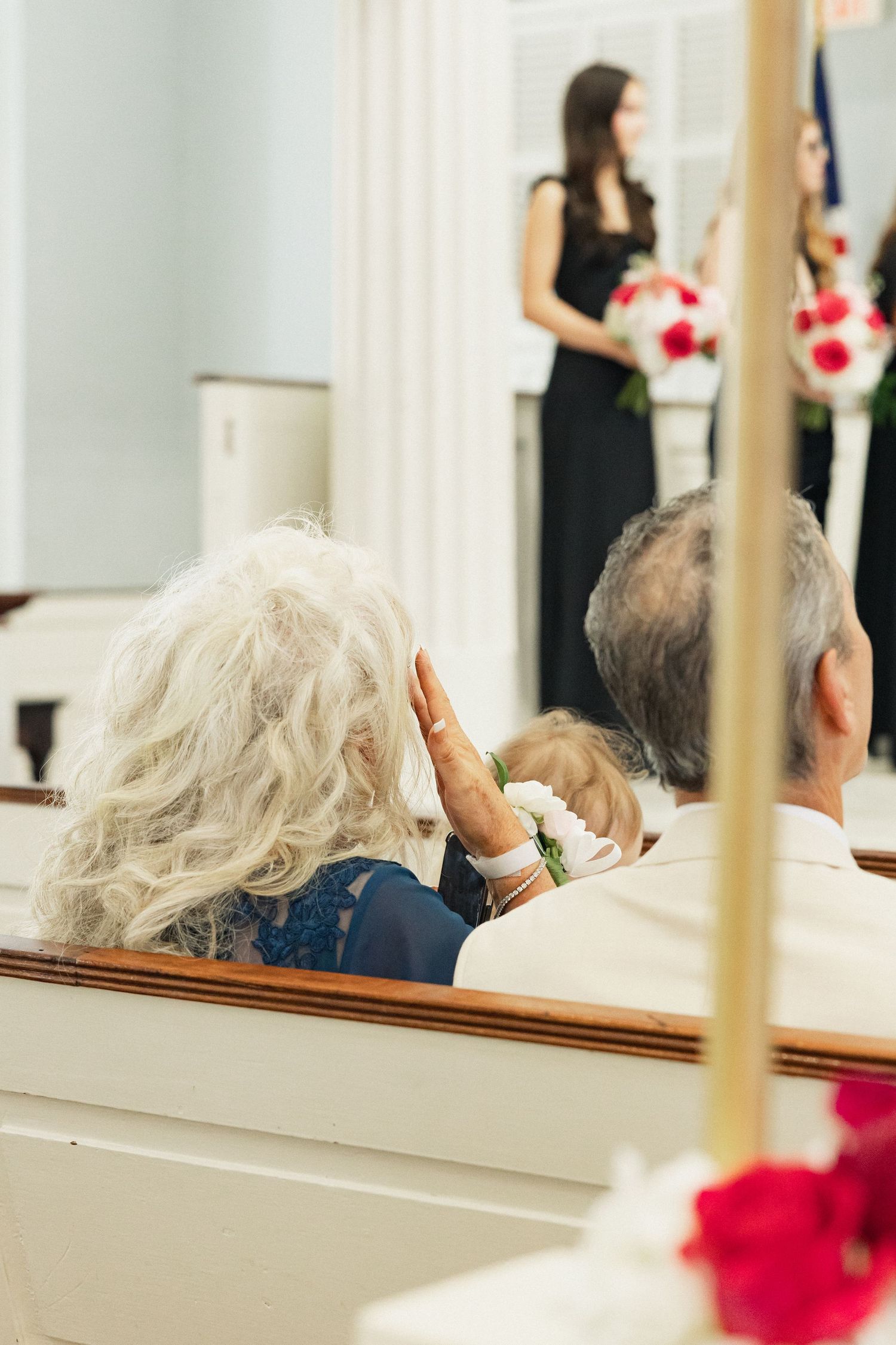 Wedding guests seated in church pews watch a bridesmaid in black holding a red and white floral arrangement at the altar.