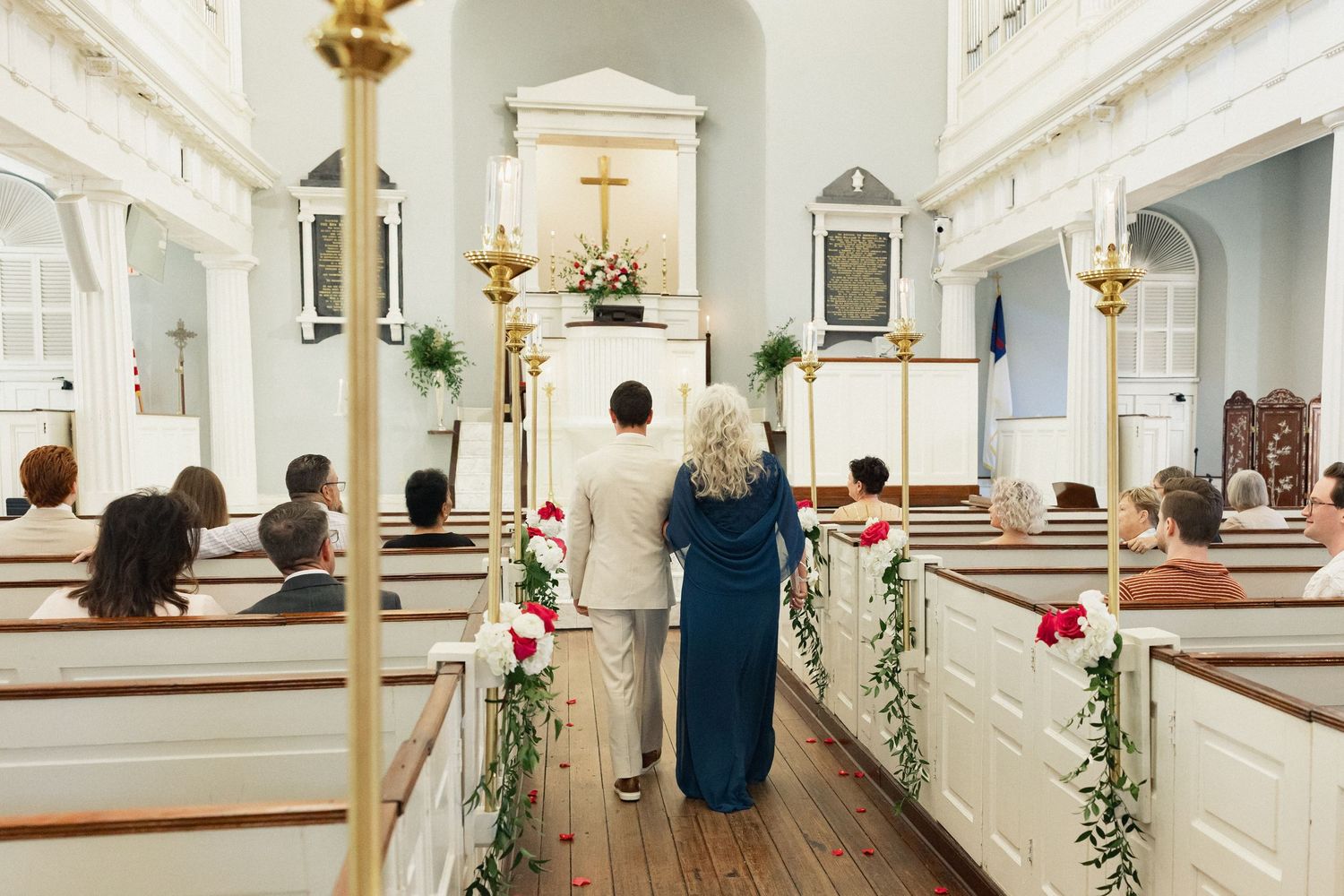 Couple walking down church aisle decorated with red roses and greenery during a wedding ceremony.