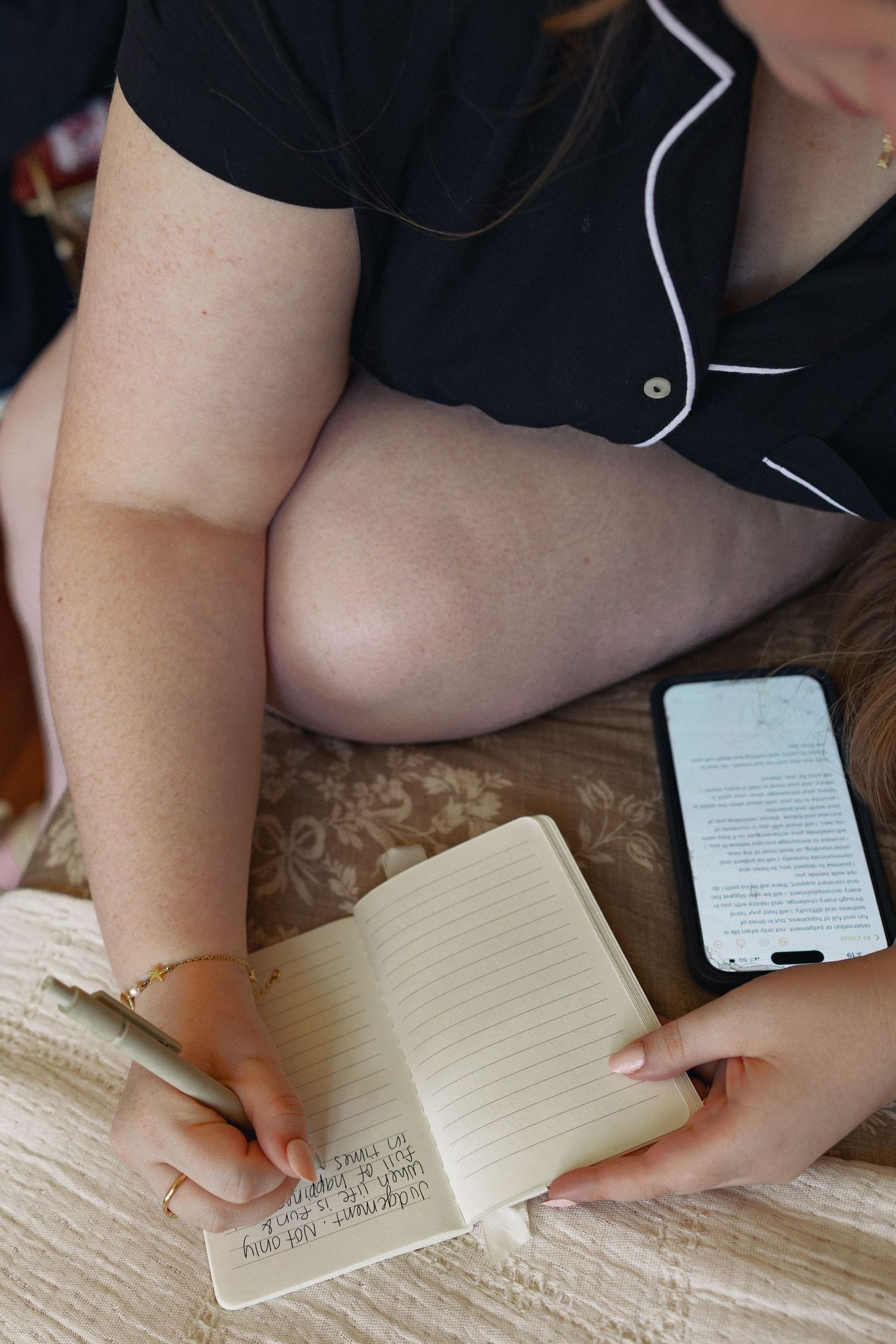 Woman in black pajamas writing in a small notebook beside a smartphone on a floral surface.