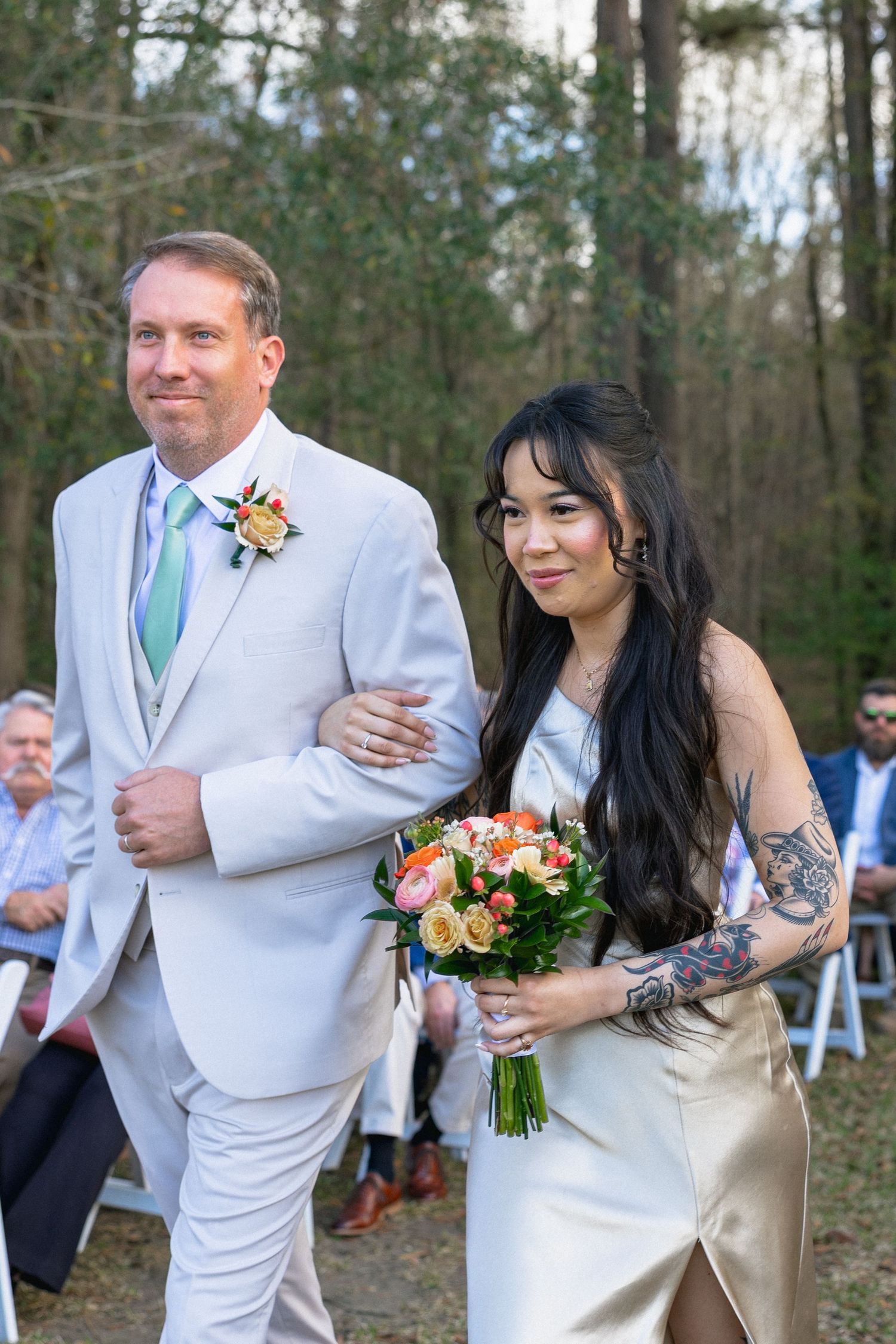A groomsman in light gray suit walks a tattooed bridesmaid in champagne dress carrying colorful bouquet down wedding aisle.