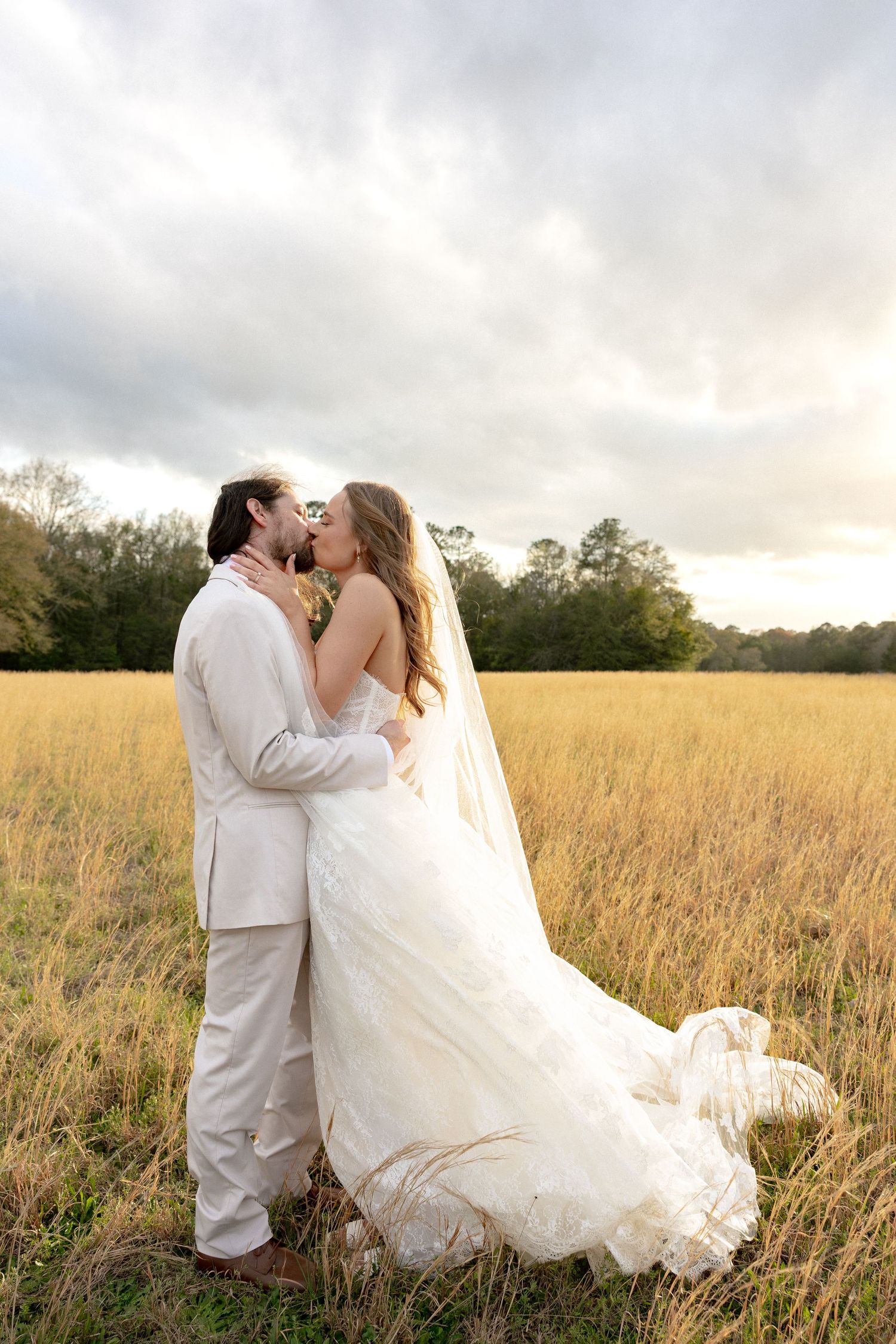 Bride and groom share a kiss in a golden grass field under dramatic cloudy skies.
