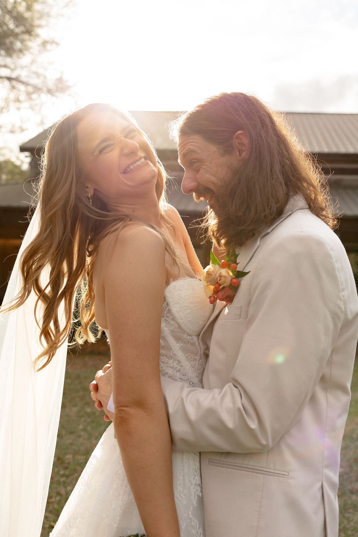 Laughing bride and groom sharing a joyful moment outdoors in golden sunlight, groom in beige suit with boutonniere.